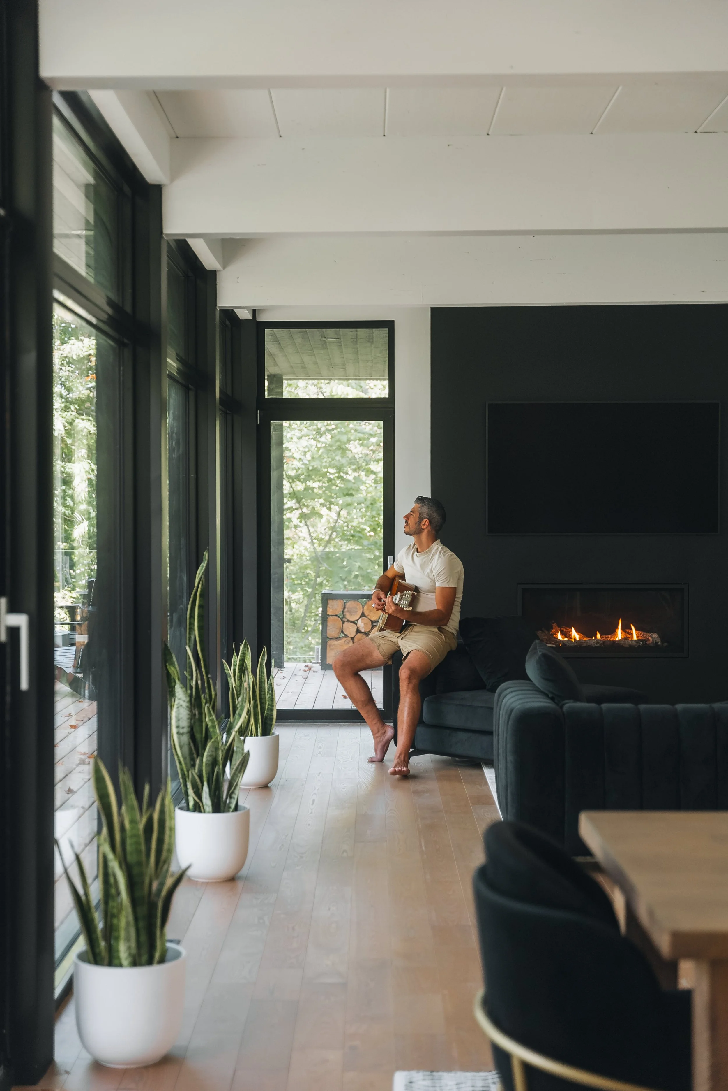 A man playing guitar on a couch in a modern living room with large windows, a fireplace, and potted plants.