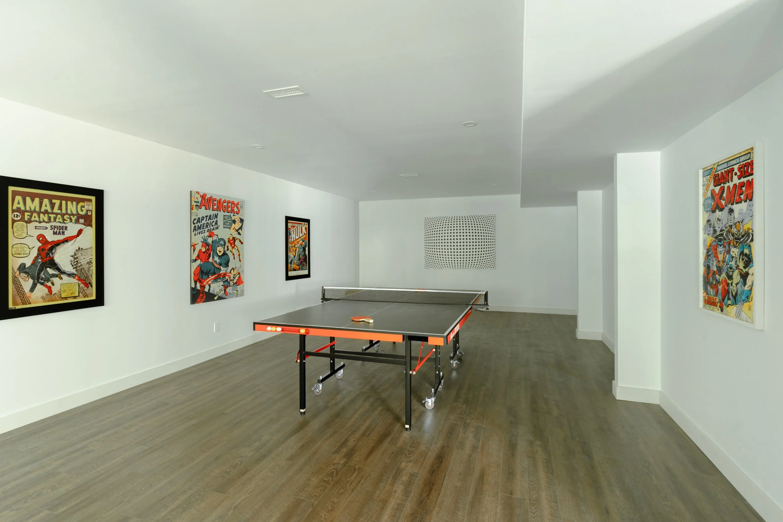 A modern room with a ping pong table in the center and comic book themed posters on the walls, featuring characters from Spider-Man, Avengers, and Hulk. The room has hardwood flooring and is brightly lit.