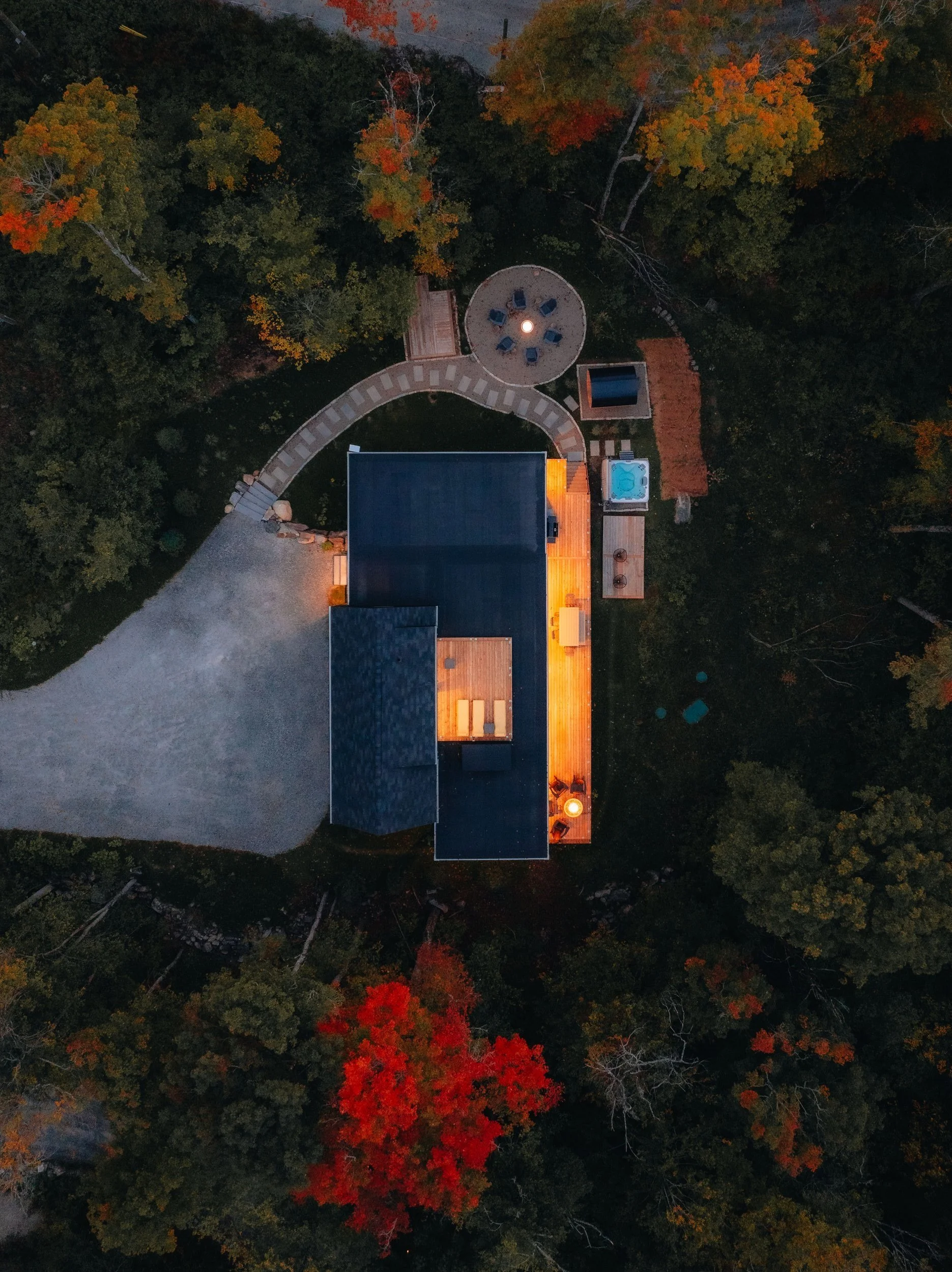 Aerial view of a modern house with patio lighting, surrounded by trees with autumn foliage. Includes a circular seating area, pathway, and a small pool or hot tub. Illuminated outdoor areas create a warm ambiance.