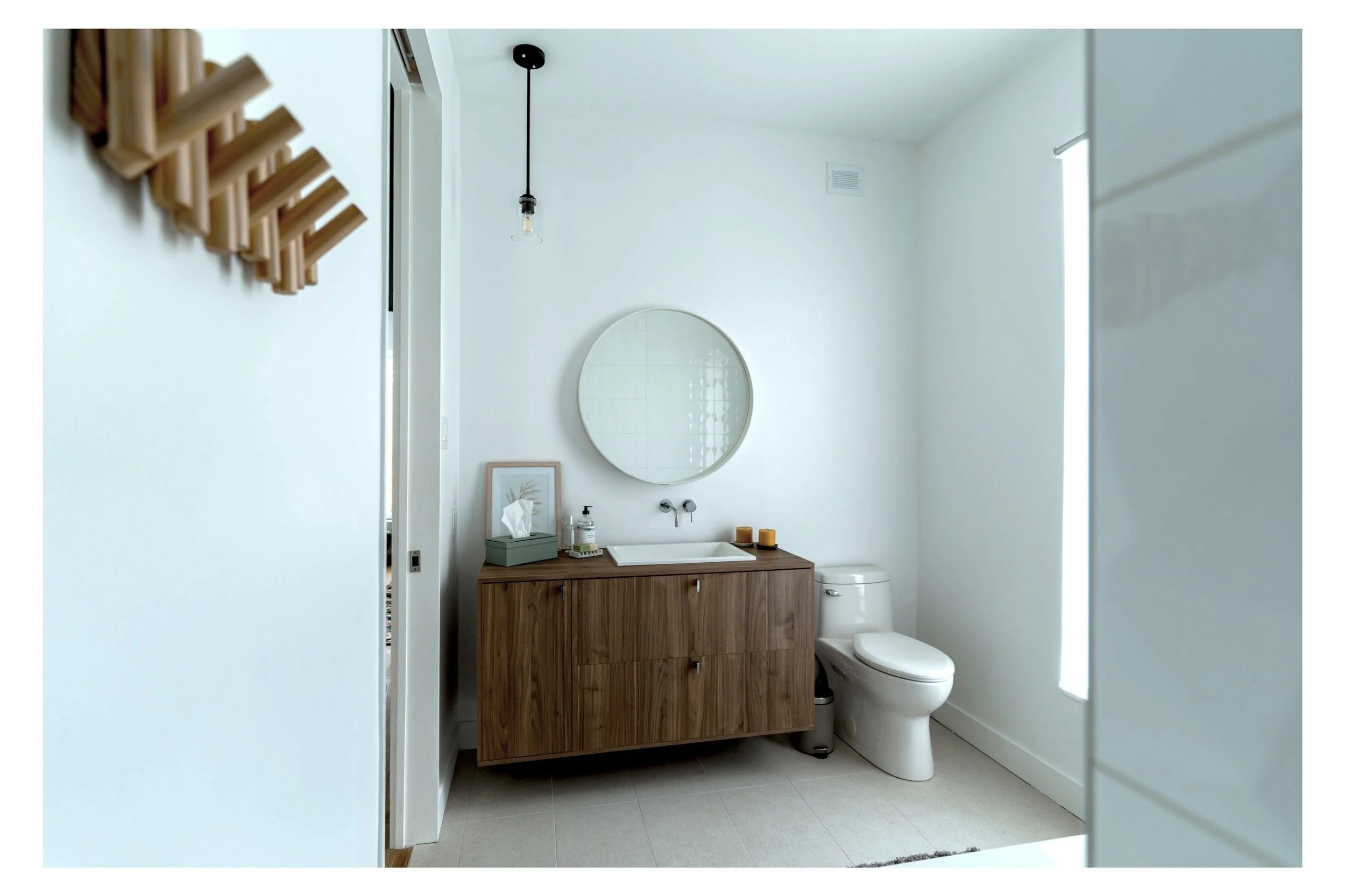 Modern minimalist bathroom with wooden vanity, round mirror, and toilet; features wall-mounted faucet, hanging light fixture, and decorative elements.