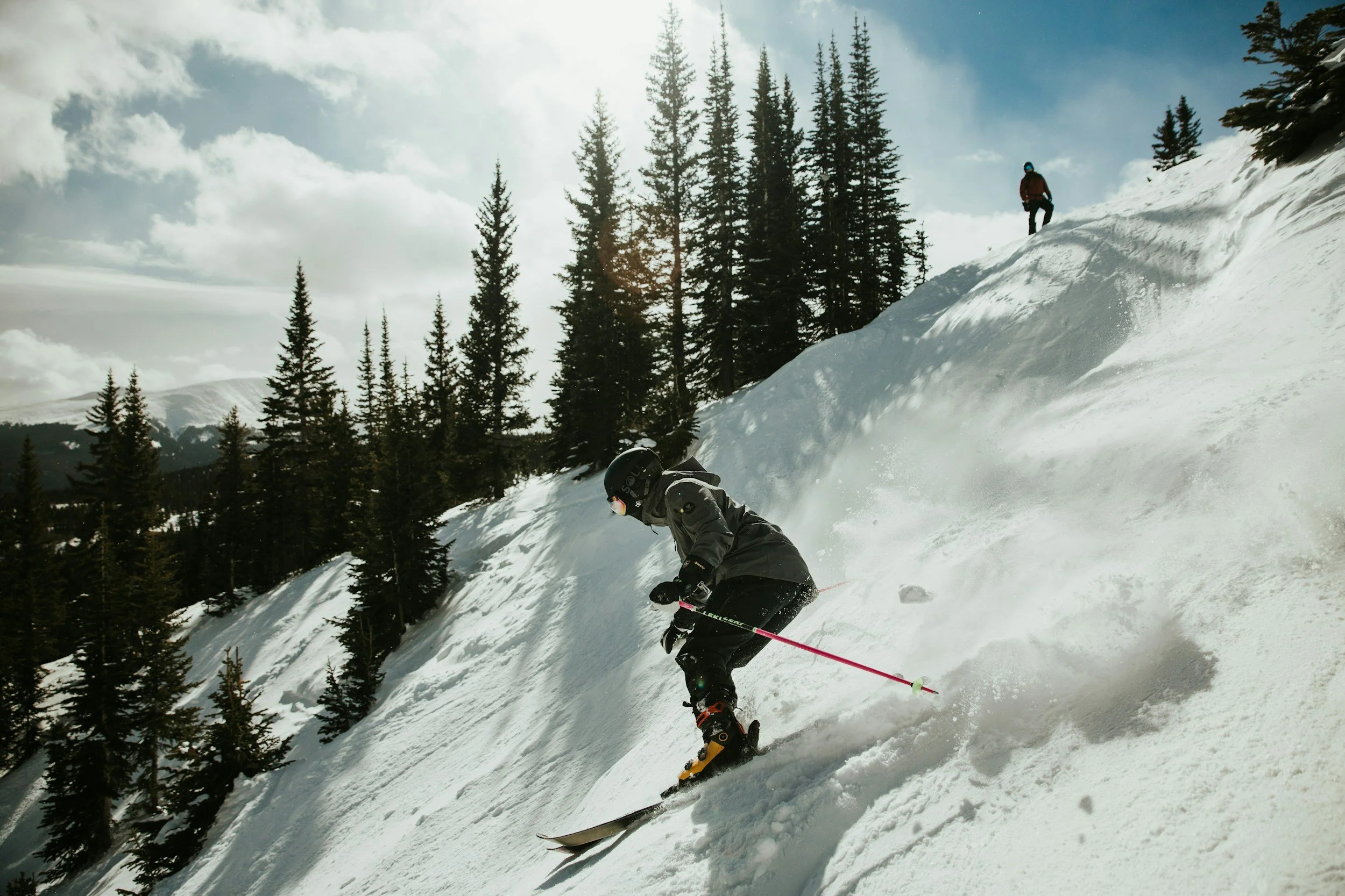 Skiers going downhill on a snowy mountain slope with trees and a sunny sky in the background.