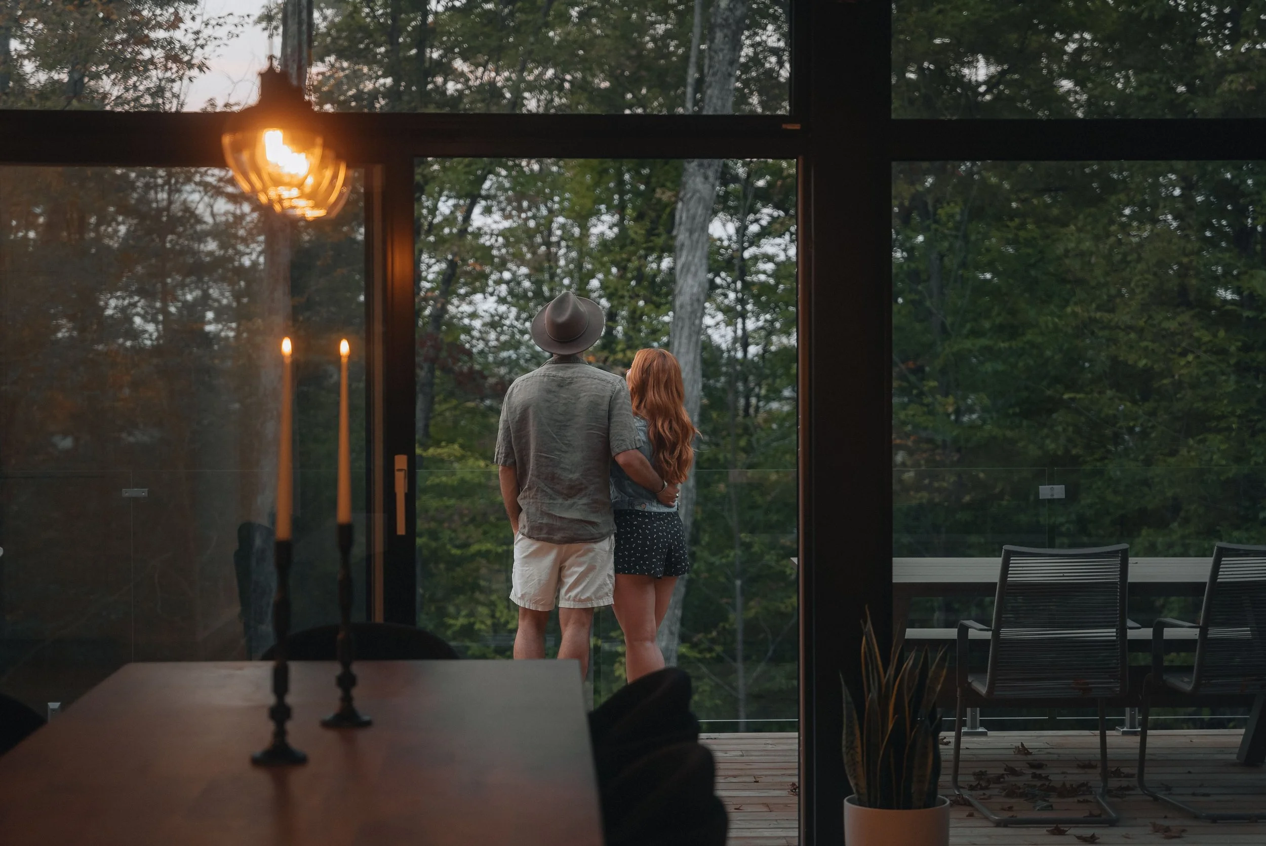 A couple stands on a balcony overlooking a forest, viewed through glass doors from inside a dimly lit room with candles on a table.