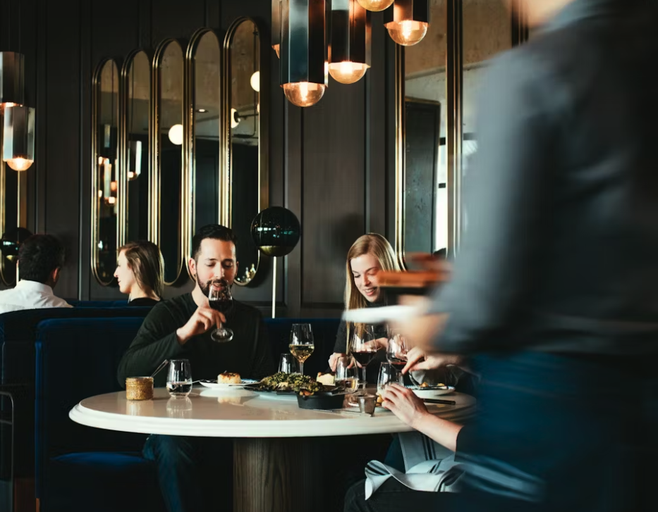People dining at a restaurant, with a man drinking wine and a woman eating, while a server walks by, modern decor with mirrors and lights in the background.