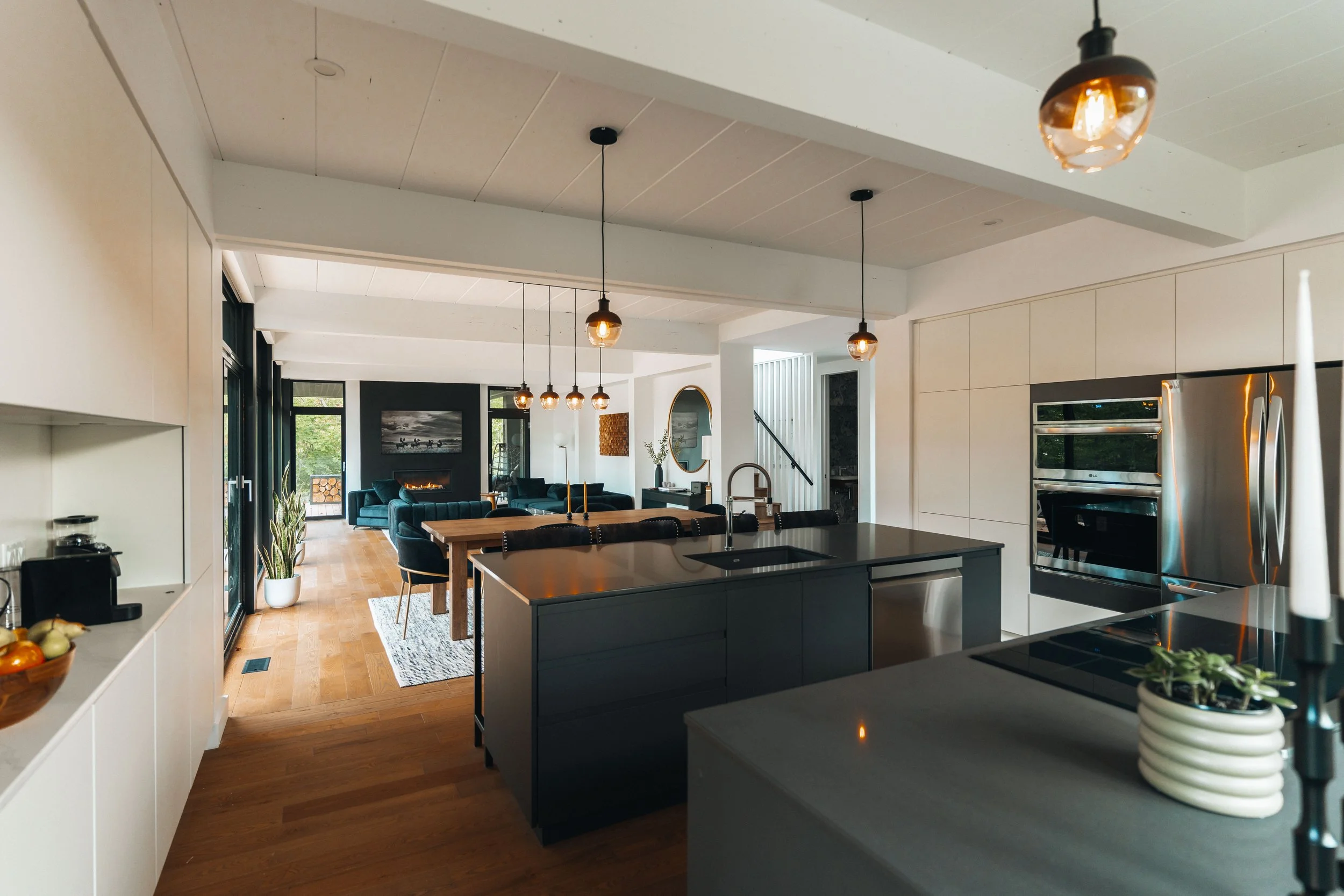Modern open-plan kitchen and living room with sleek cabinetry, stainless steel appliances, a large island with pendant lighting, wooden flooring, and a seating area with dark sofas and fireplace.