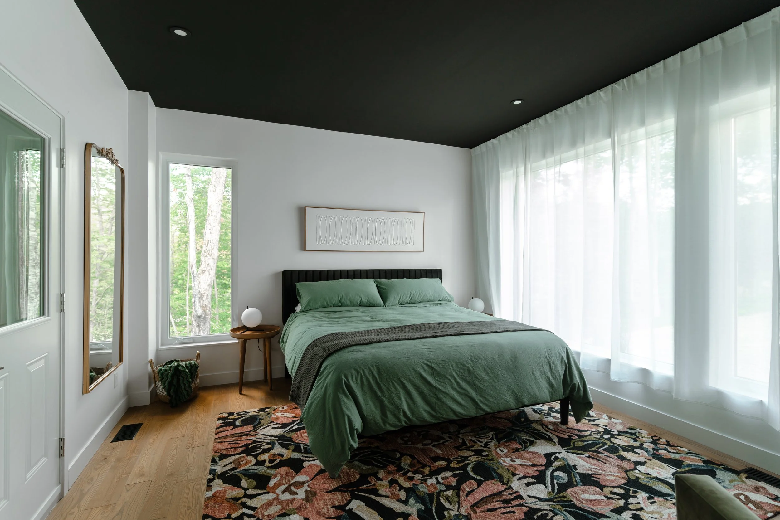 Modern bedroom with green bedding, floral rug, and large windows letting in natural light.
