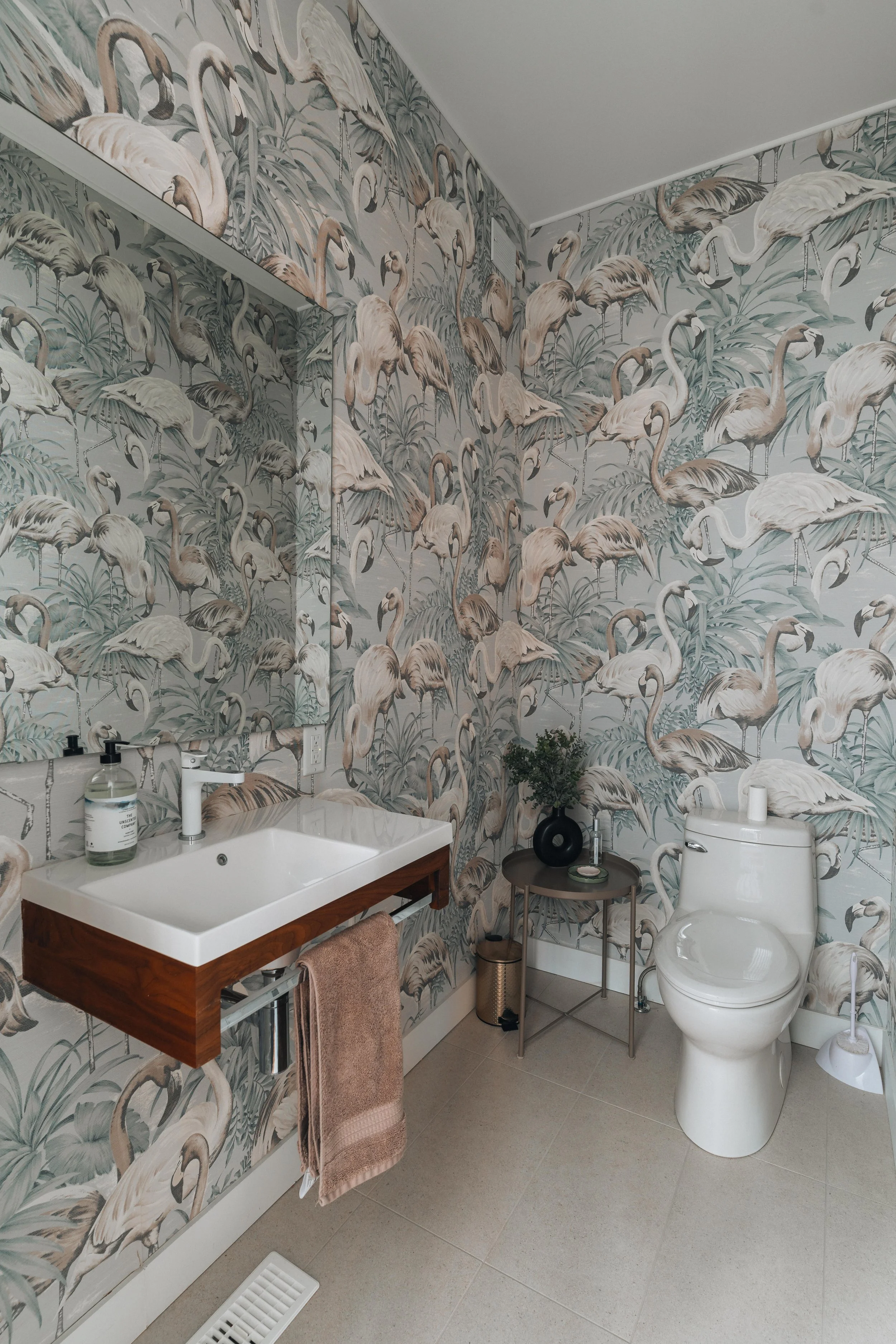 Bathroom with flamingo patterned wallpaper, a white sink with a wooden base, a brown towel, a round side table with decor, and a white toilet.