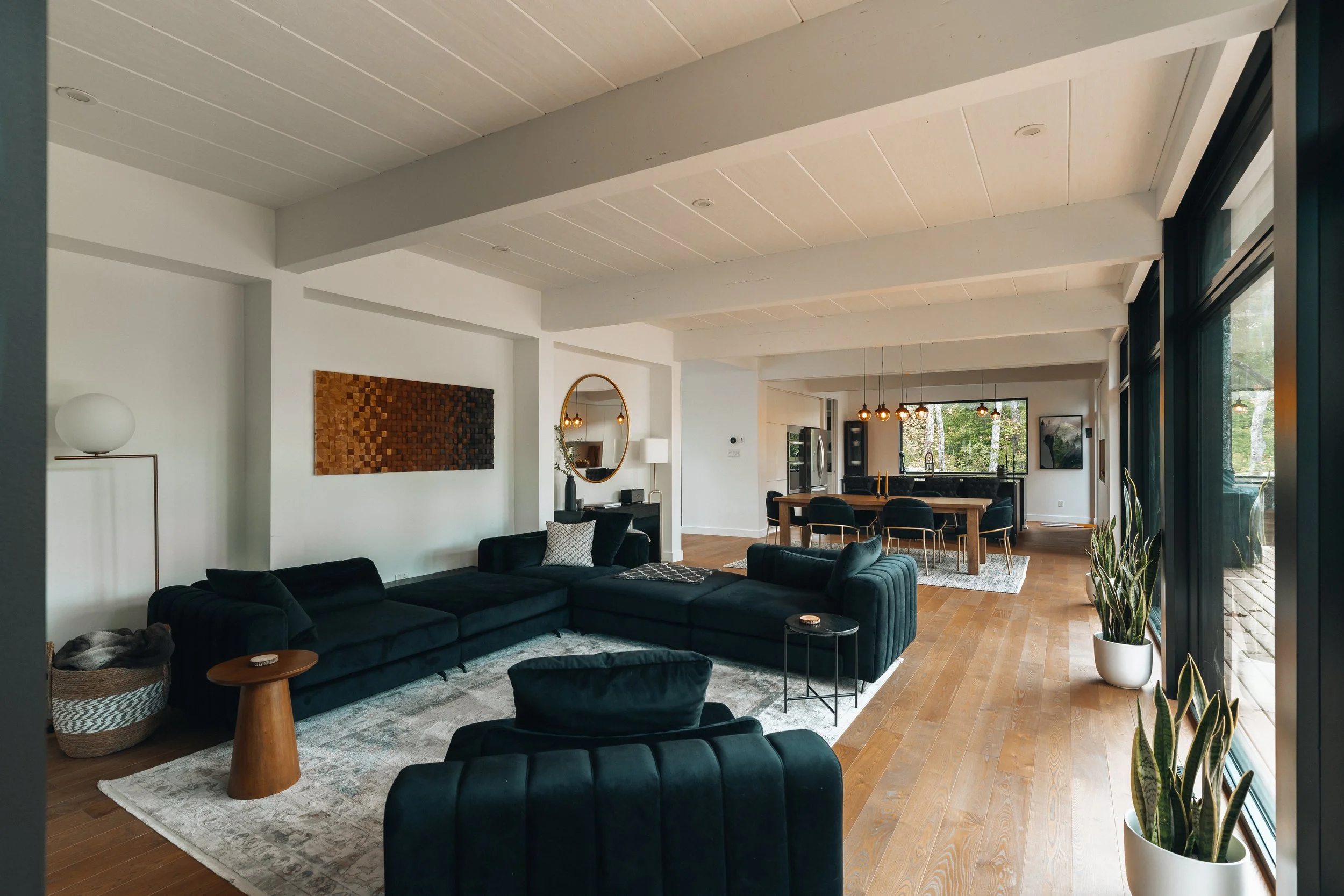 Modern living room with dark green velvet sectional sofa, wooden coffee tables, art on white walls, a dining area with a large table, hanging lights, houseplants, and large windows showing a wooded view.
