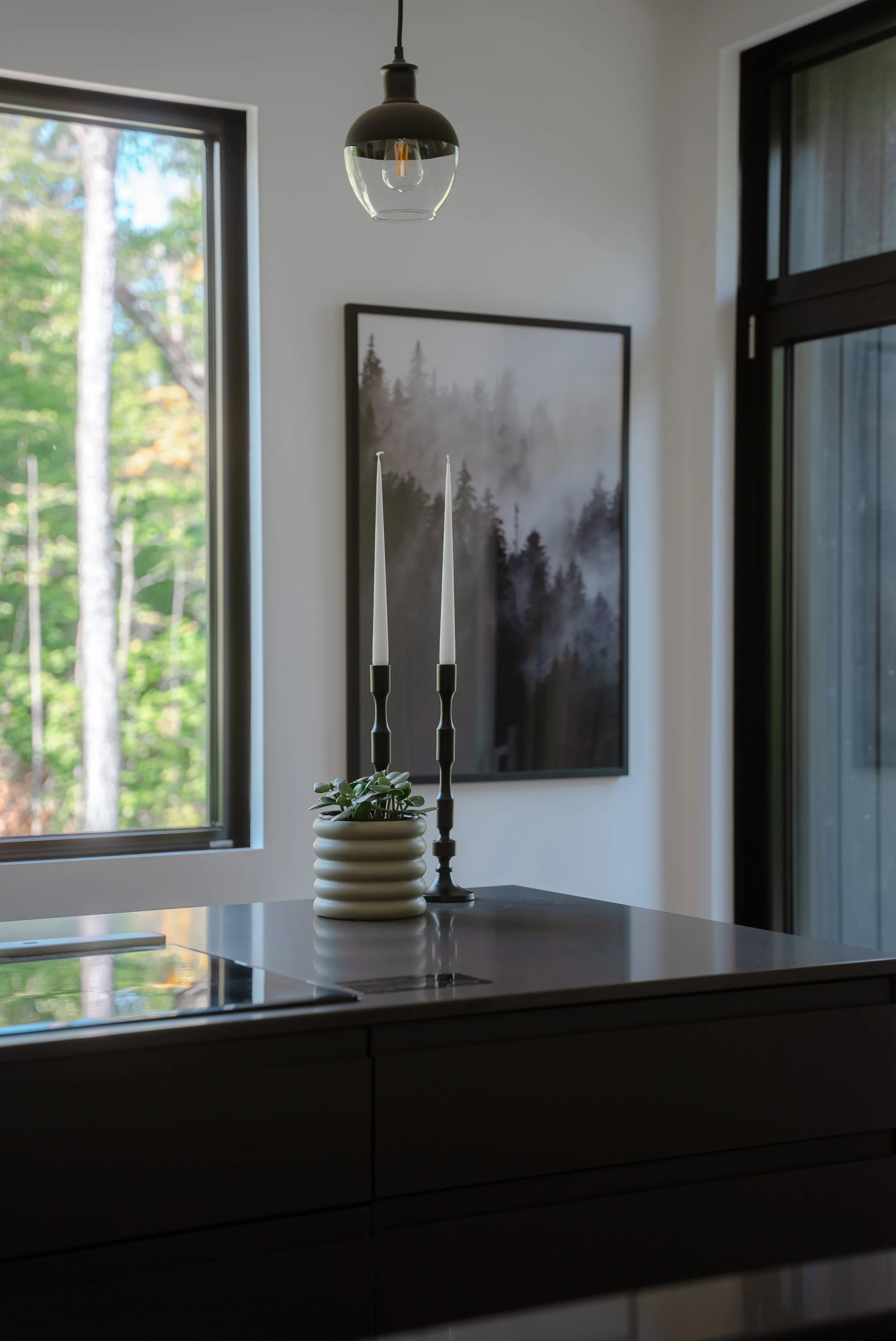 Modern kitchen interior with a pendant light, two tall white candles in black holders, a small potted plant on a countertop, and a framed forest landscape painting on the wall.