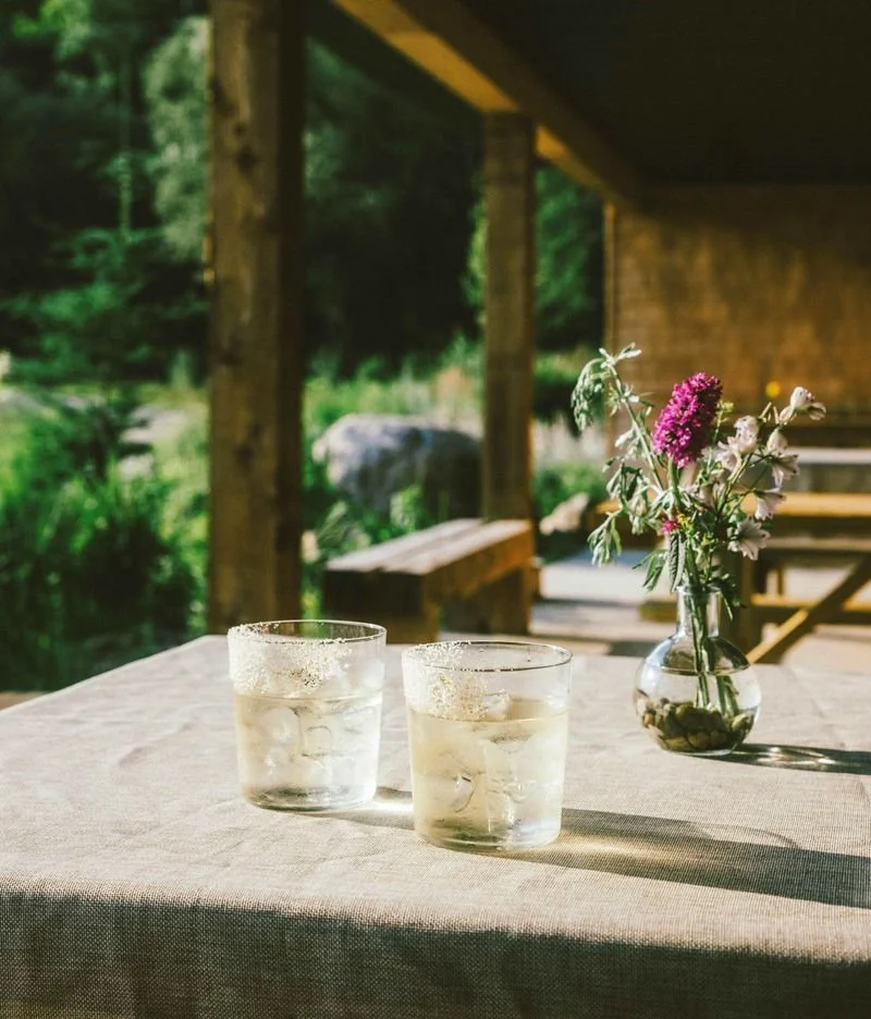 Two glasses of iced drinks on a table with a flower vase outdoors.