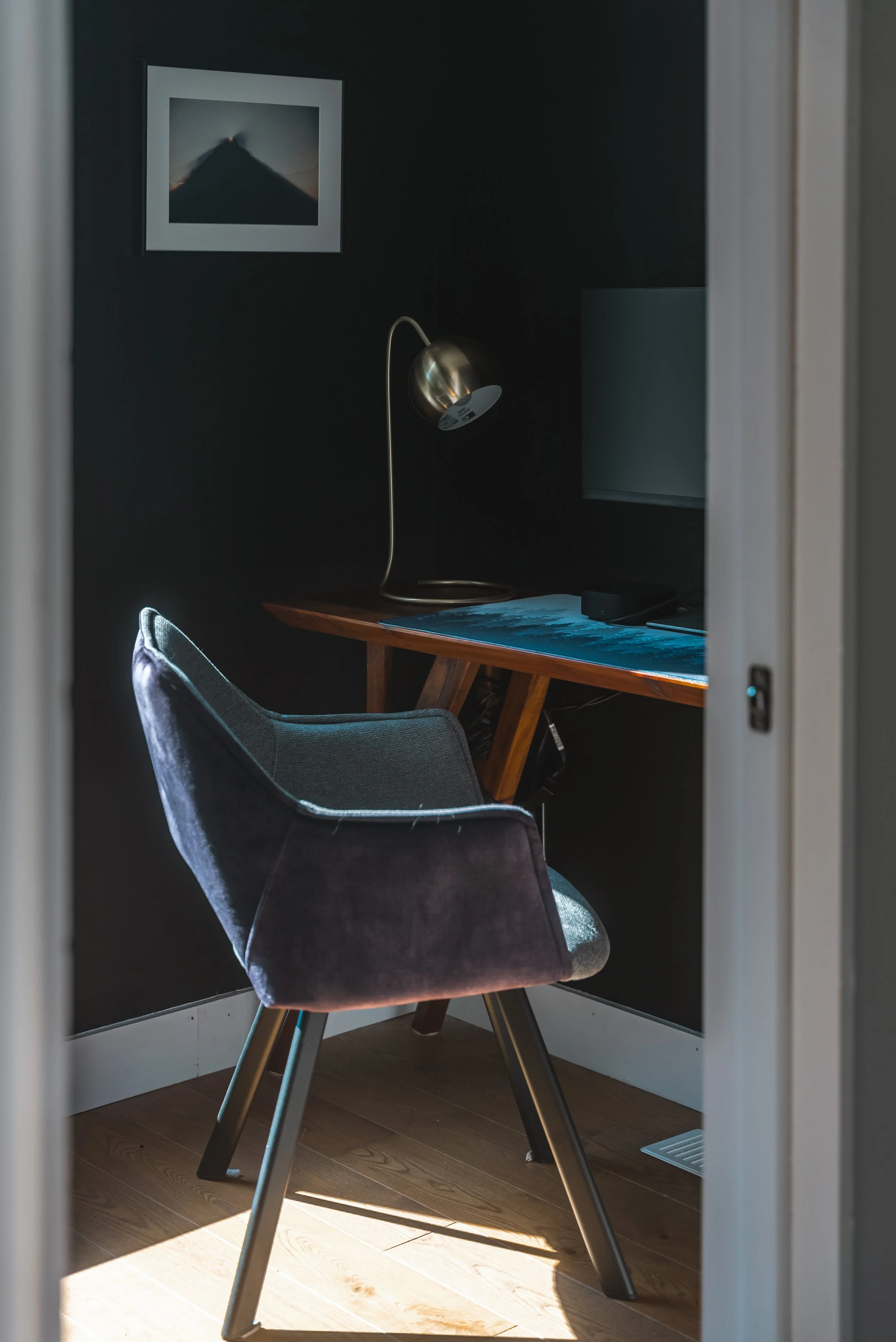 Minimalist home office with black walls, a wooden desk, gray chair, brass desk lamp, and framed artwork.