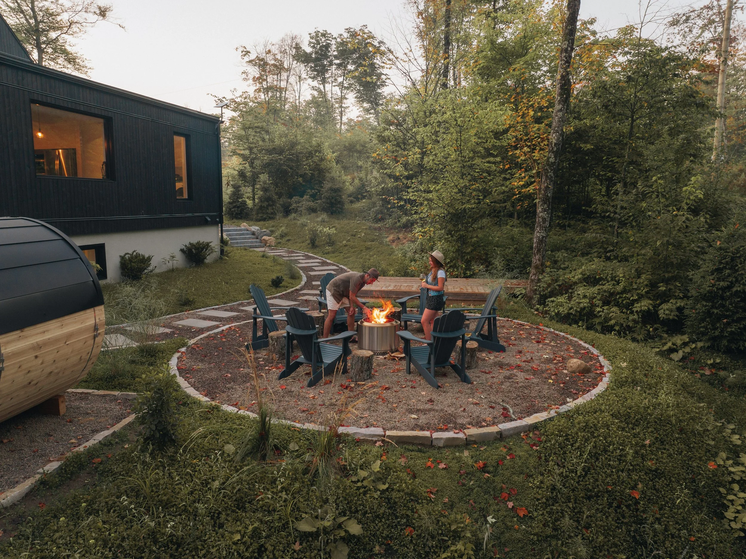 Two people by a fire pit, surrounded by Adirondack chairs, in a forested backyard near a modern house with large windows.
