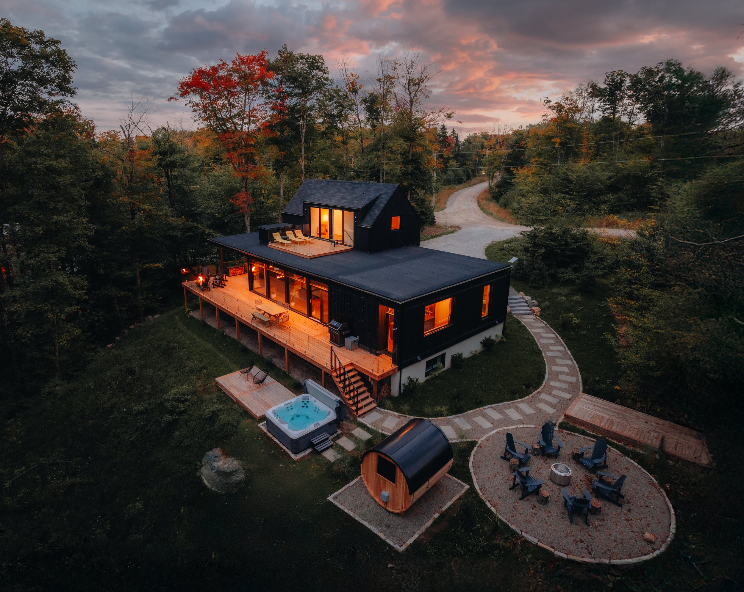 Aerial view of a modern, two-story house in a wooded area at sunset, featuring a wooden deck, hot tub, and outdoor seating area with a fire pit. The surrounding trees have autumn colors, and a winding path leads to the house.