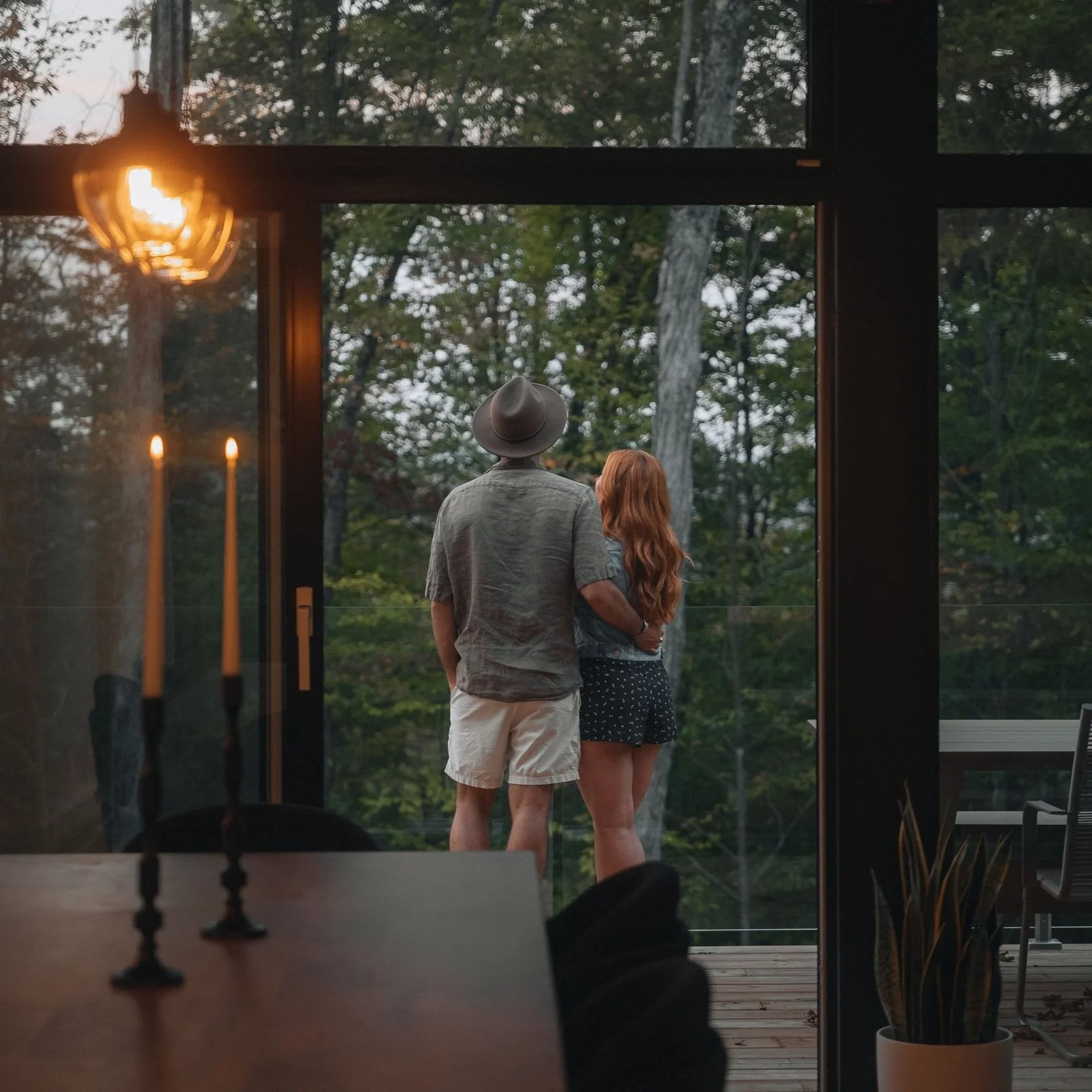 A couple stands on a balcony overlooking a wooded area during dusk, with warm indoor lighting and lit candles visible inside the home.