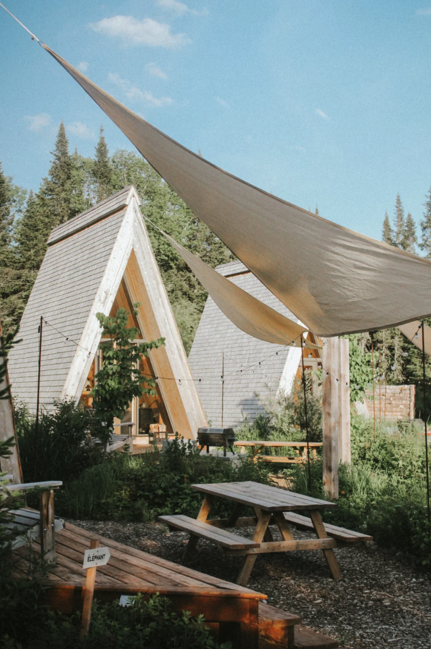 A-frame cabins in a wooded area with picnic tables and sun sails, creating a rustic outdoor setting.