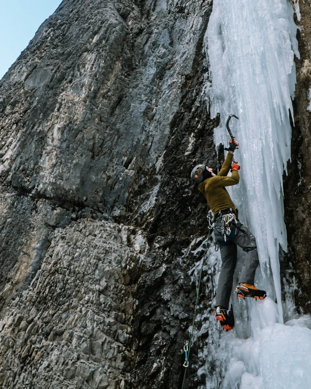 Person ice climbing with ice axes on a frozen waterfall, wearing climbing gear and crampons, against a rocky cliff.