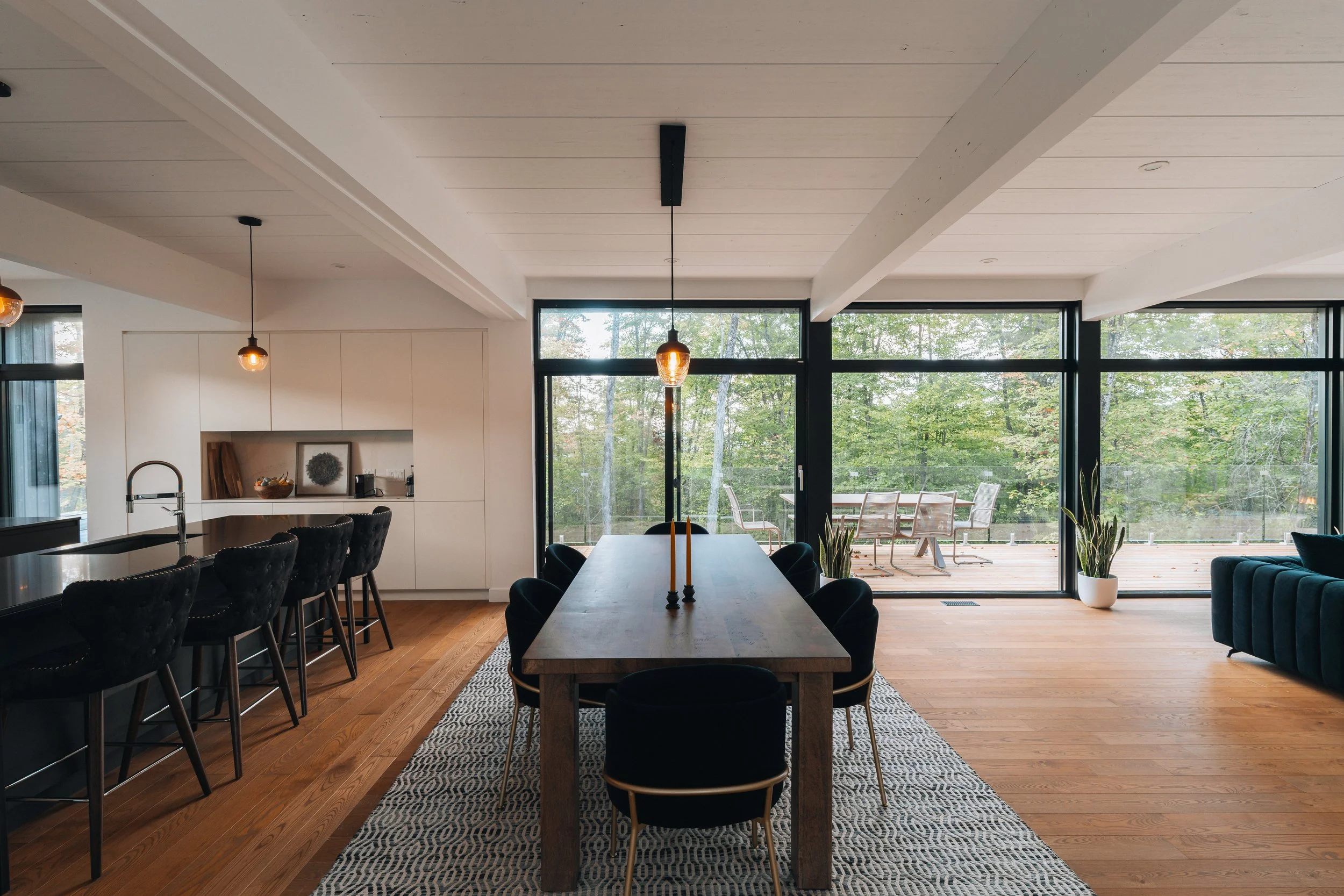 Modern open-concept dining and kitchen area with wooden floors, a wooden dining table with black chairs, pendant lighting, and large glass doors leading to an outdoor patio.