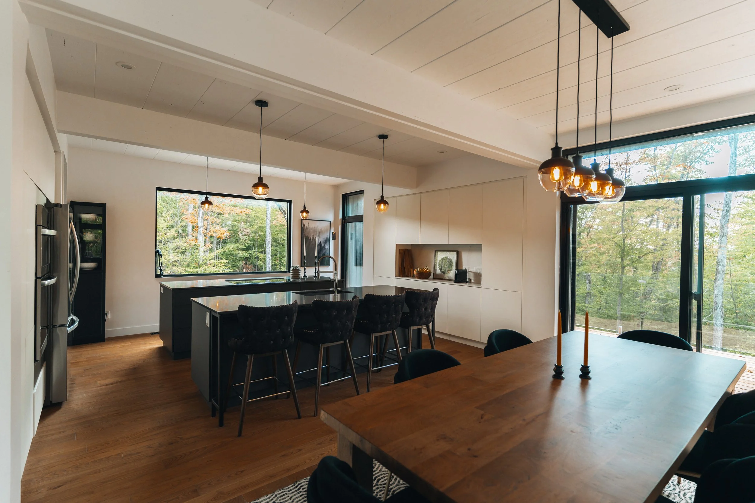 Modern kitchen and dining area with large windows, wooden flooring, and pendant lights. Features a kitchen island with barstools, and a dining table with candles.