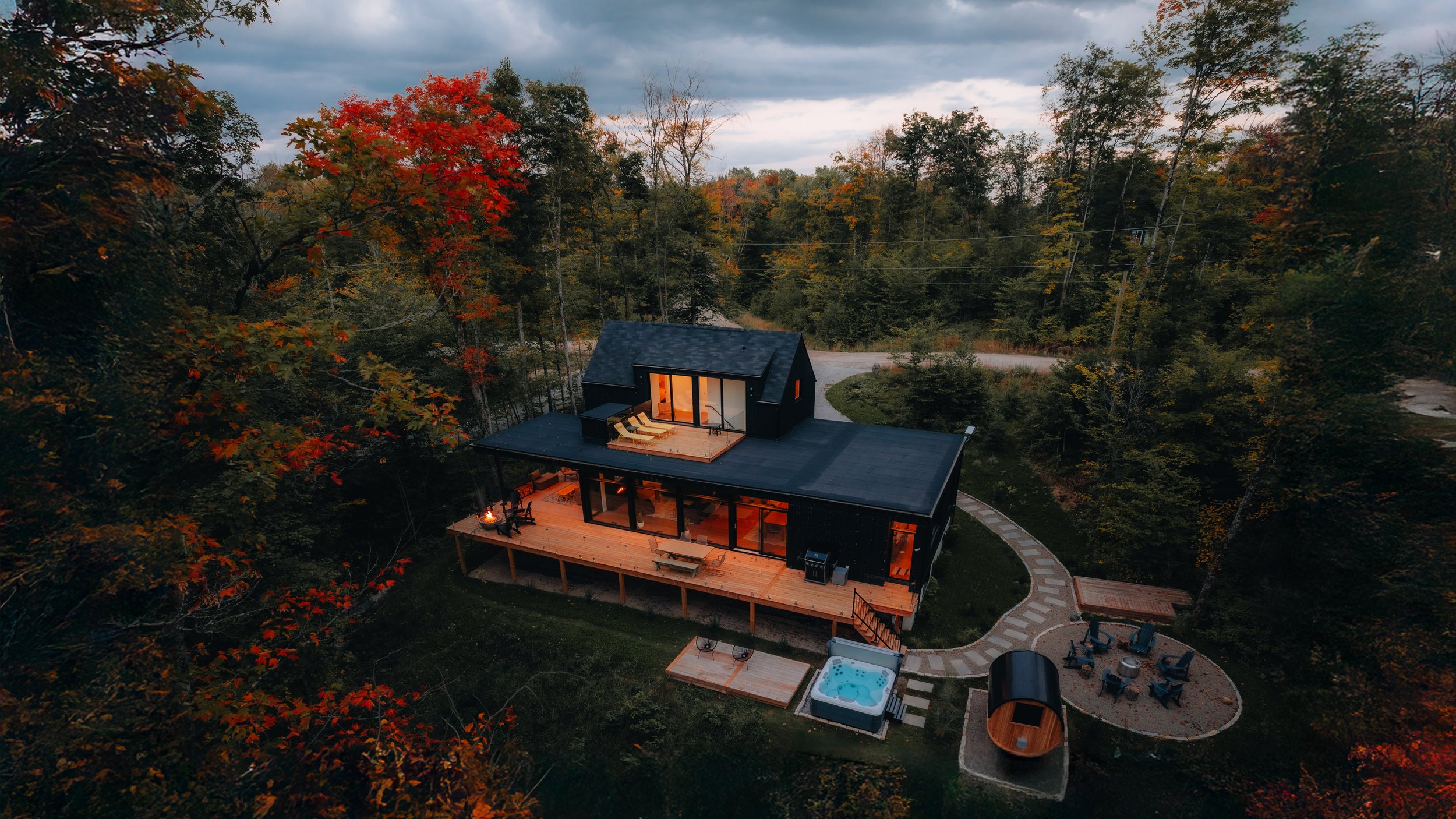 Modern cabin in a forest with autumn foliage, featuring a deck, lounge area, and hot tub.