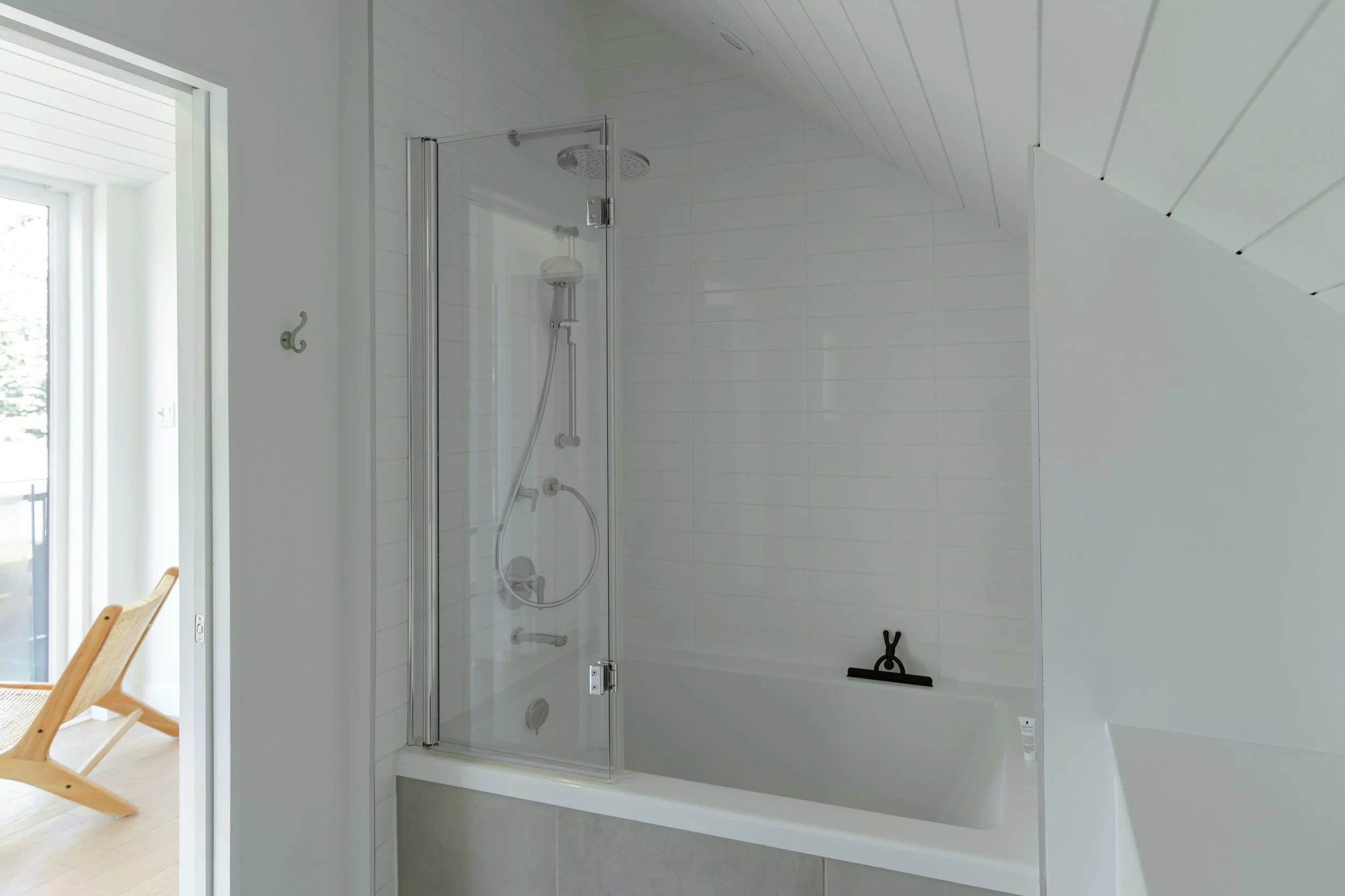 Modern bathroom with a glass-enclosed shower and white bathtub, minimalistic design, light gray tile, and a wooden chair visible outside.