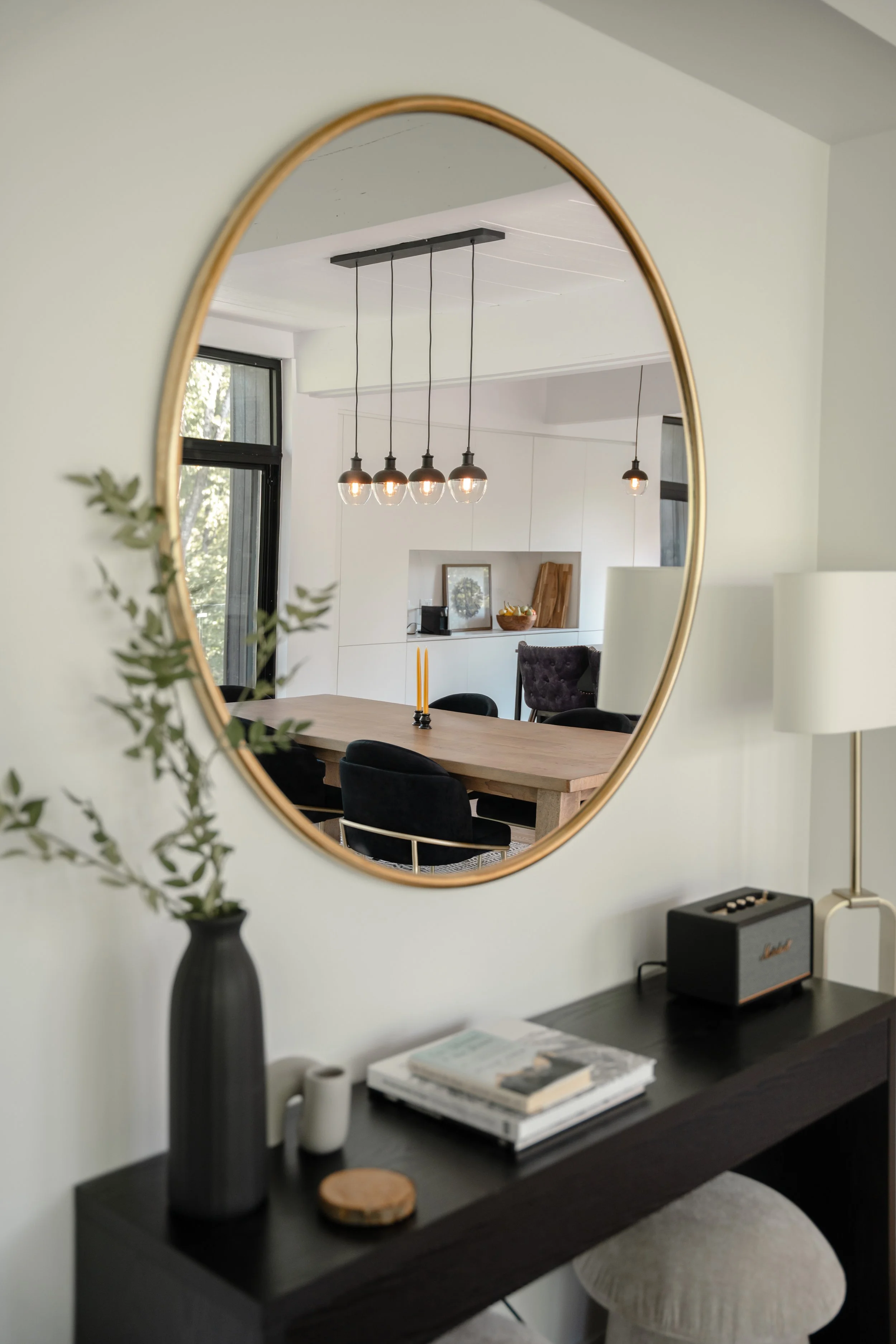 Modern dining room reflected in round mirror with black table and decor items including a vase, books, and speaker; pendant lights above dining table.