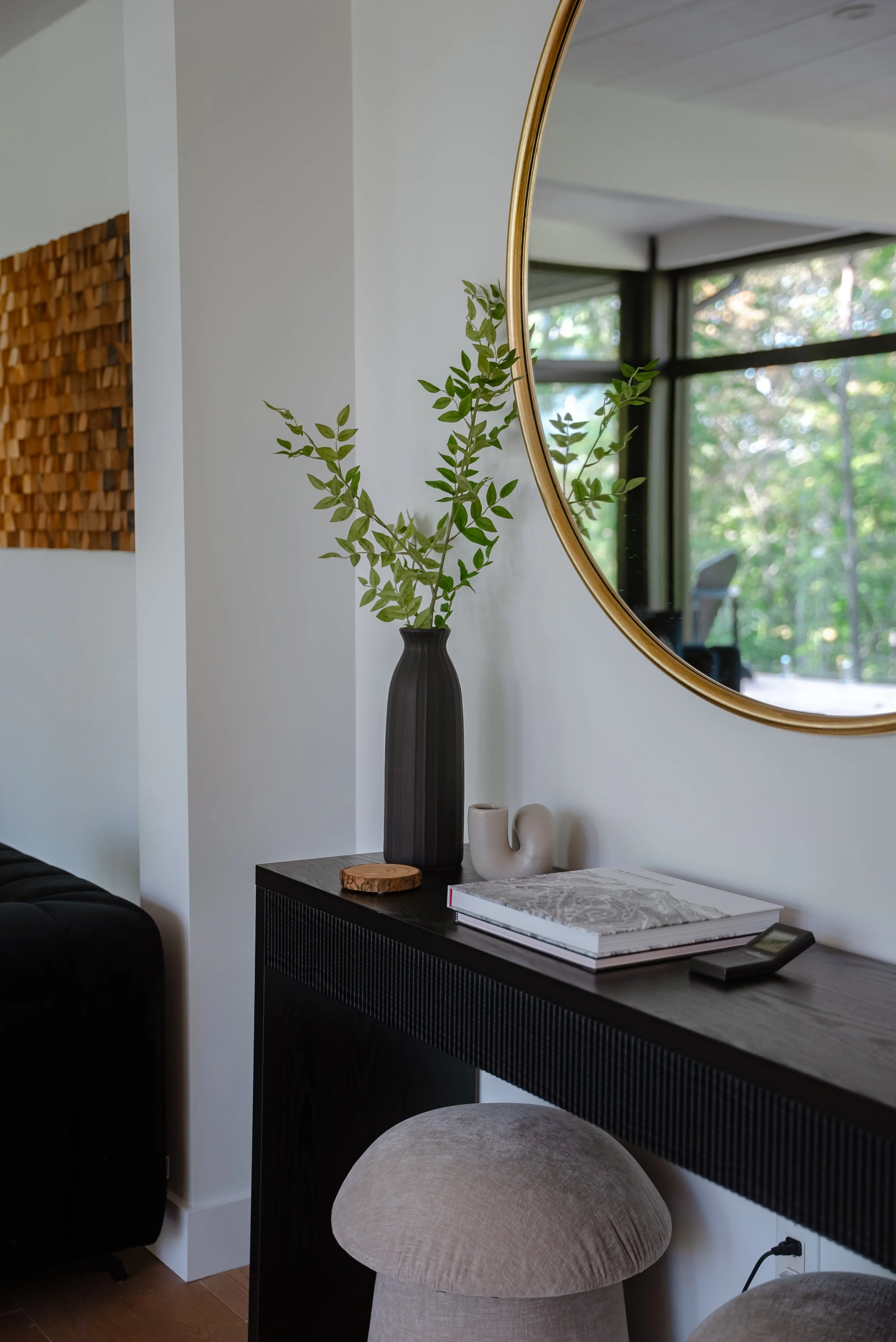 Interior design with a modern console table featuring a black vase with green foliage, books, and decorative items. A round gold-framed mirror hangs above, reflecting the bright room and large windows with garden views.