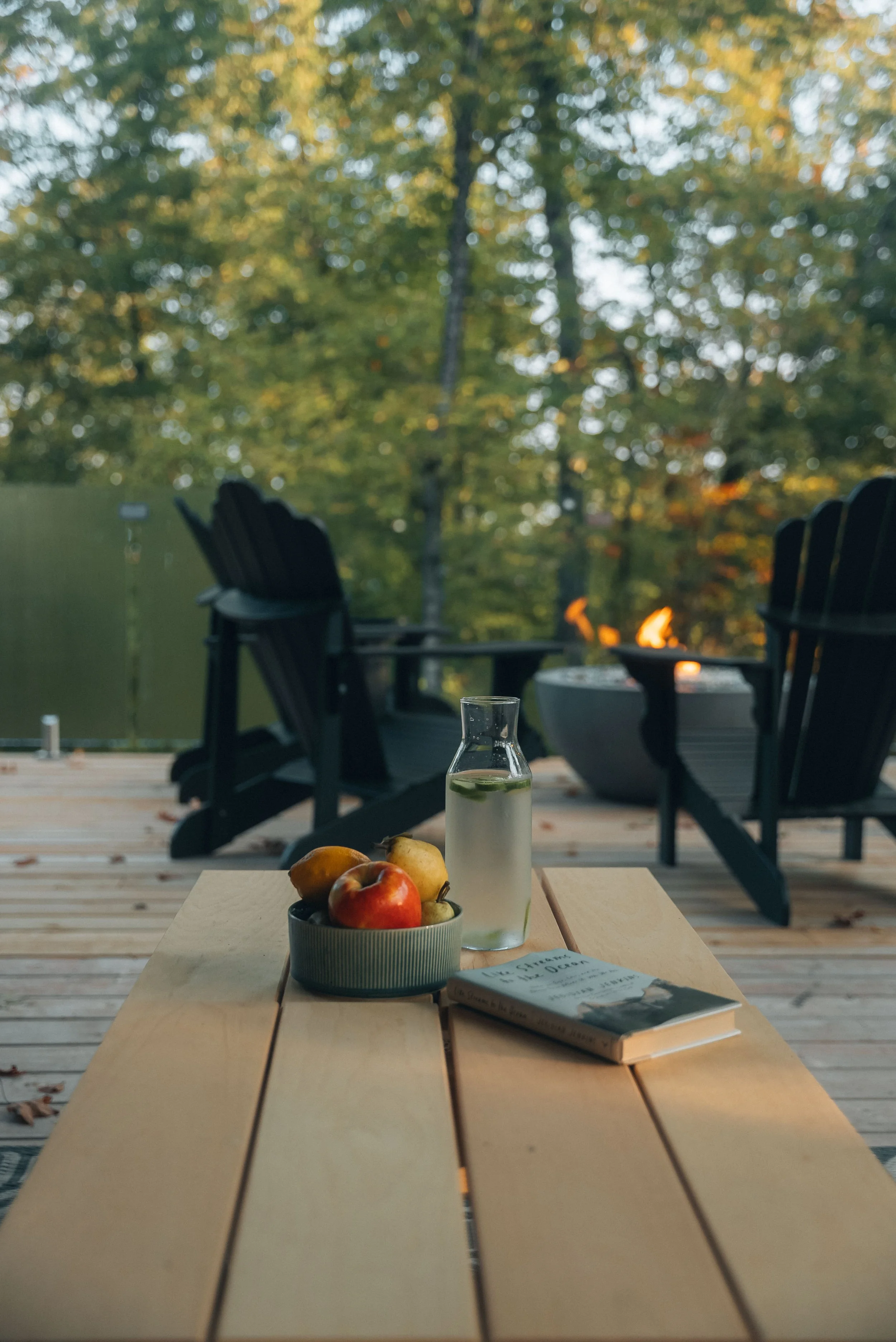 Outdoor deck scene with a wooden table, a bowl of fruits, a bottle of water, and a book, patio chairs, and a fire pit, with trees in the background.