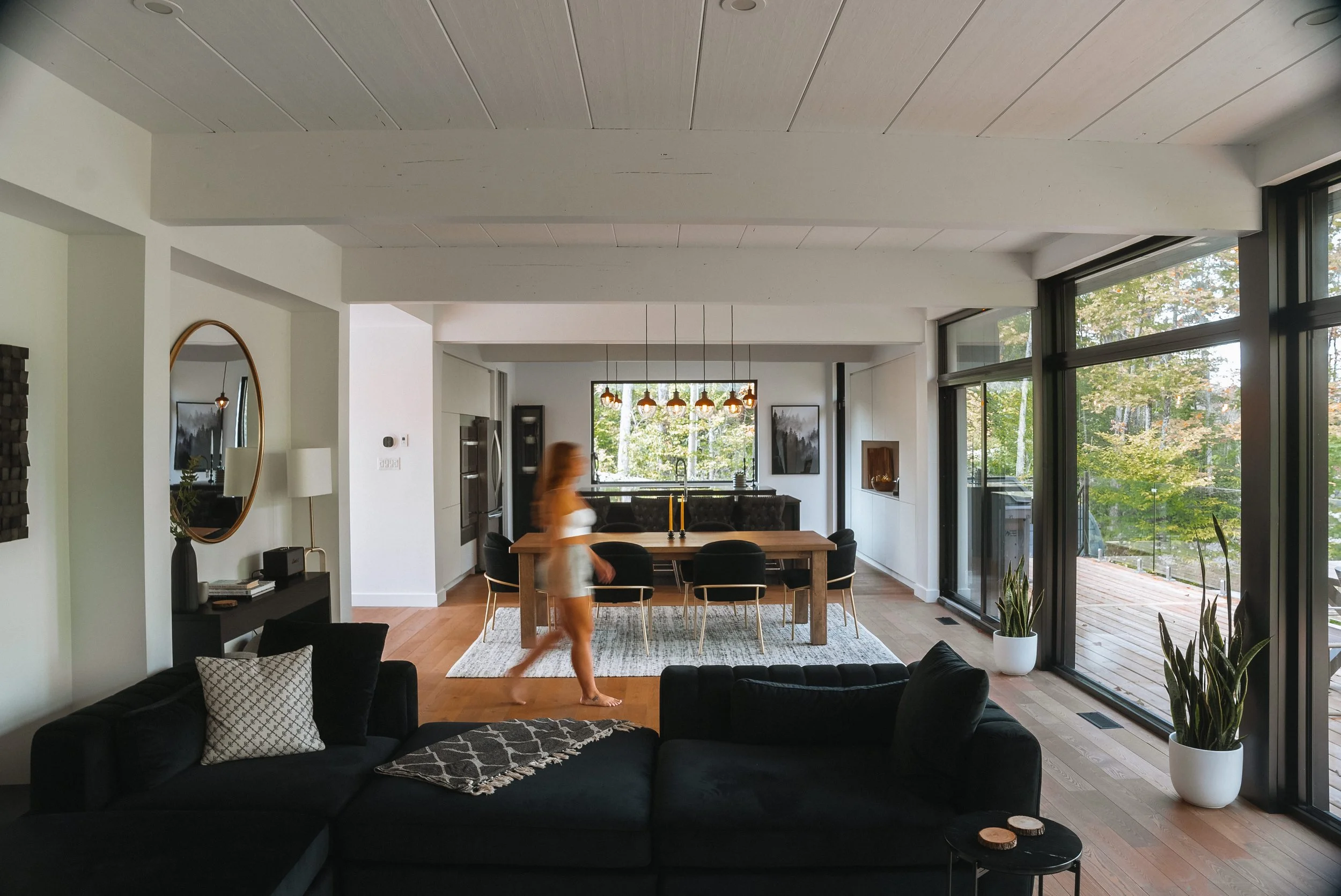 Modern living and dining room with large windows, a black sofa, wooden floor, round mirror on the wall, dining table with chairs, and indoor plants. A person is walking through the space.