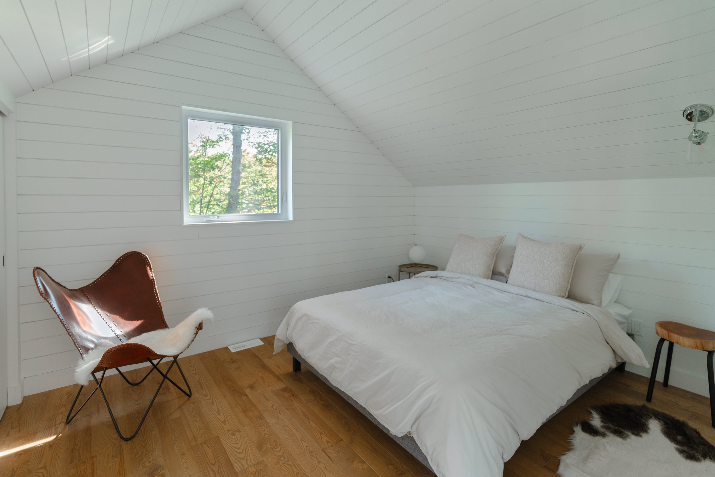 Minimalist bedroom with white walls, a bed with white bedding and beige pillows, a brown leather chair with a fur throw, and a small wooden stool on a wooden floor.