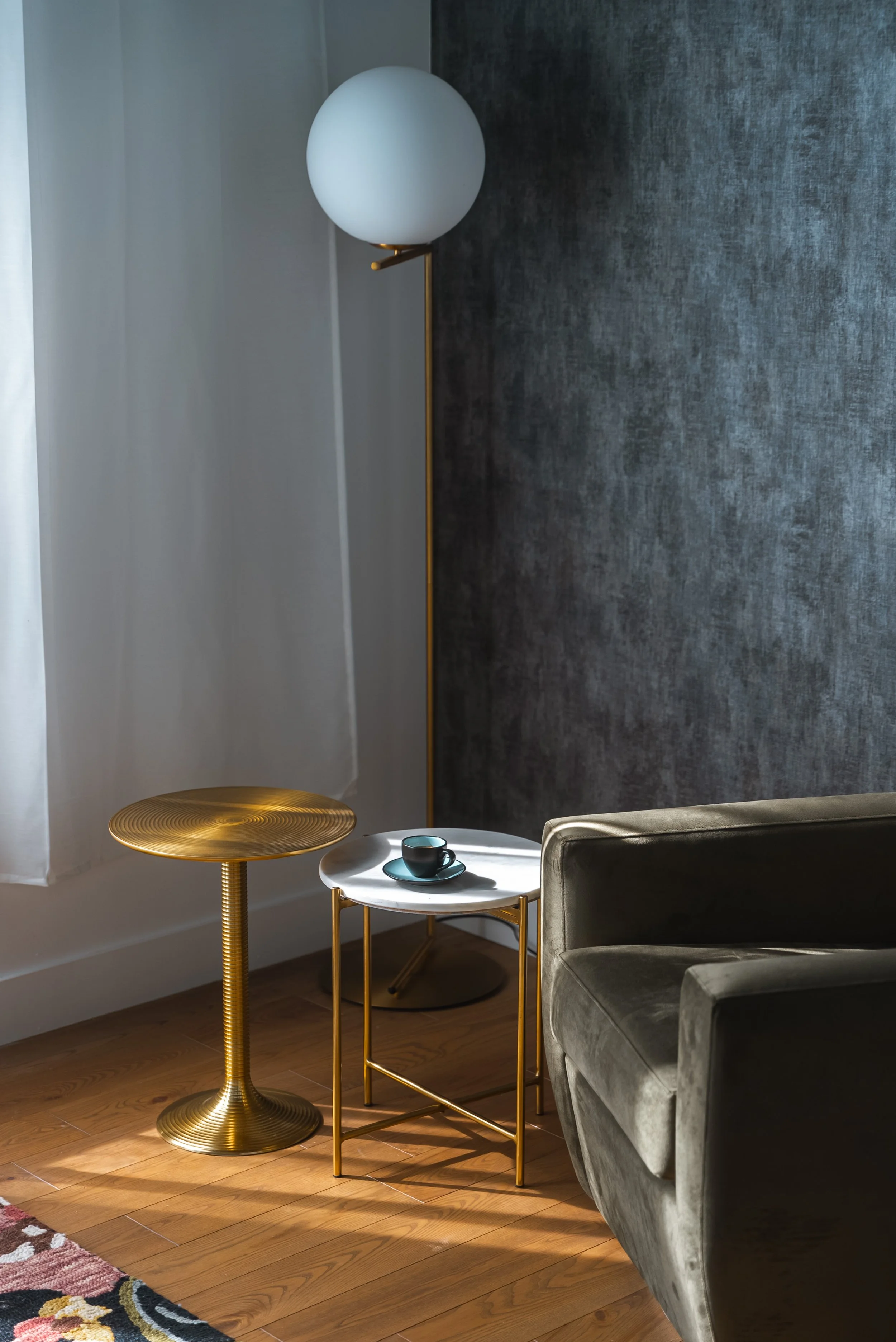 Modern living room interior featuring a green velvet armchair, a gold floor lamp with a white globe, a small round gold table, and a marble-top side table with a black cup and saucer, set against a textured gray wall and wood flooring.