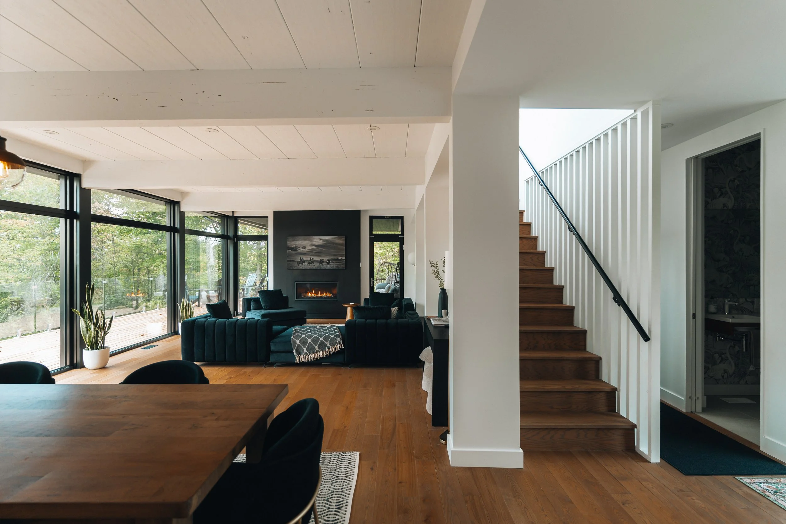 Modern living room with large windows, dark teal sofas, wooden floors, dining table, indoor plants, and a staircase. A fireplace is on the dark accent wall.