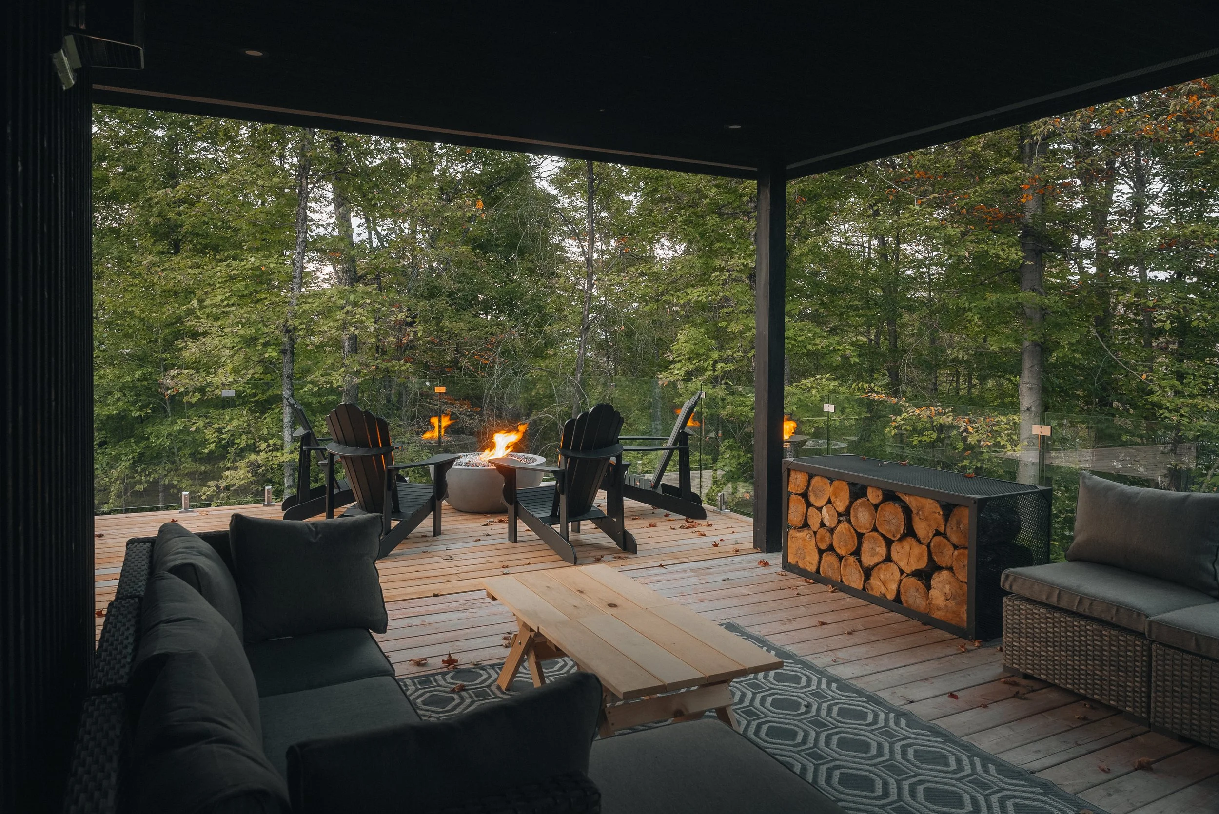 Covered patio with outdoor furniture, fire pit, stack of firewood, and forest view.