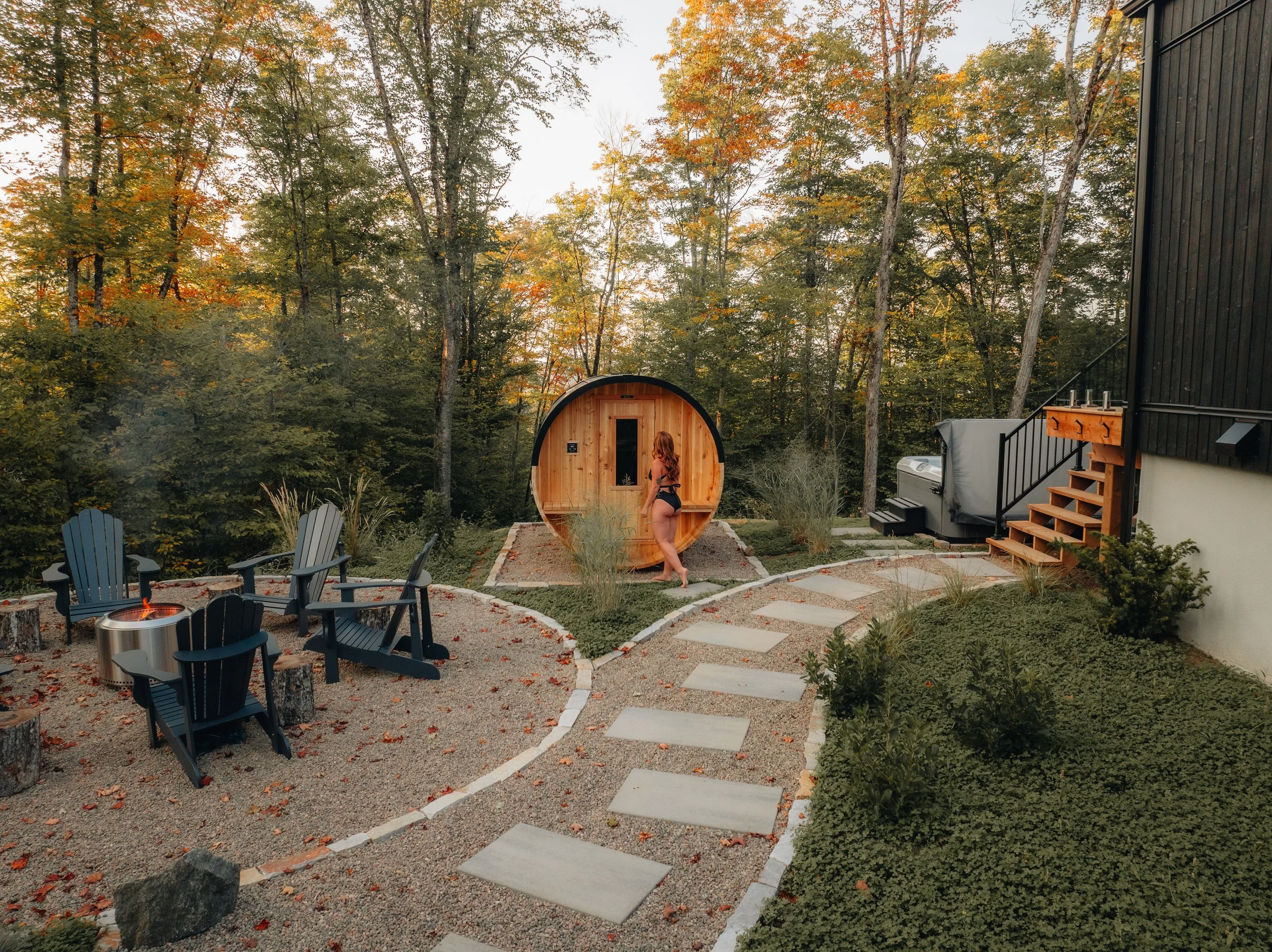 Outdoor scene with a wooden sauna, woman in swimwear, Adirondack chairs around a fire pit, surrounded by trees and a stone pathway.