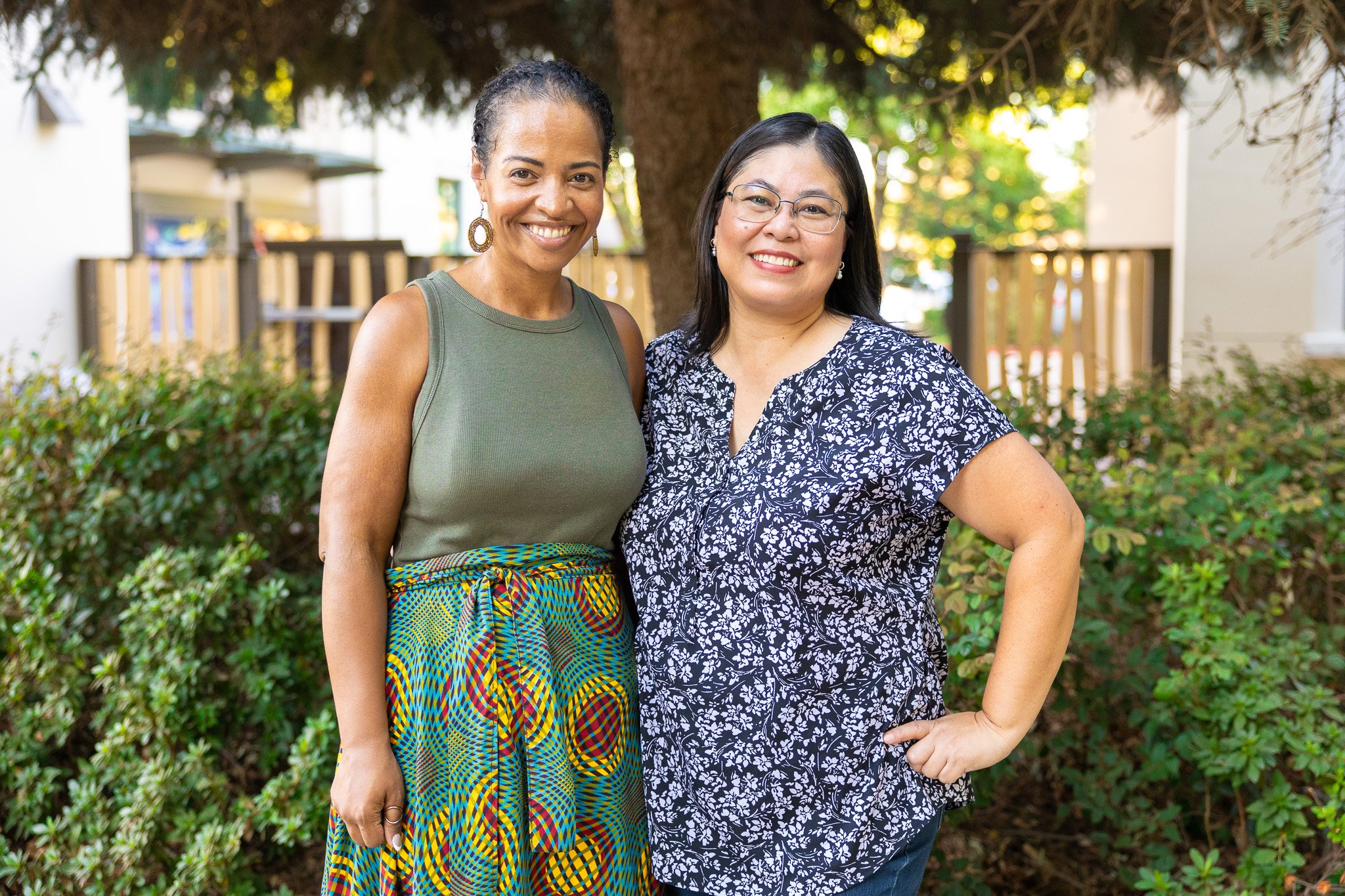 Two smiling women standing outdoors in front of a tree and bushes, one wearing a green sleeveless top and colorful patterned skirt, the other wearing glasses and a blue floral blouse.