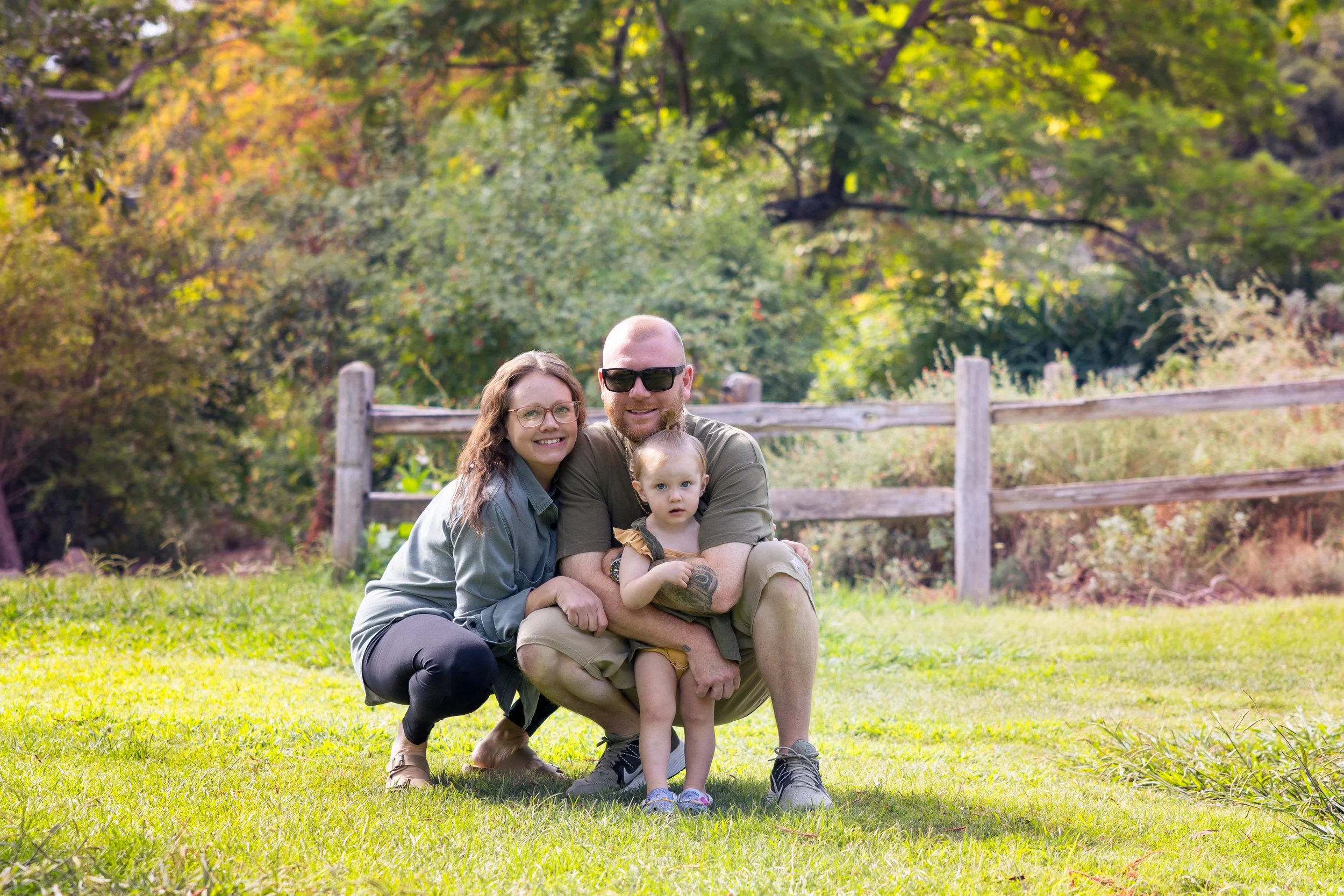 A family of three smiling and sitting on grass outdoors with a wooden fence and trees in the background.