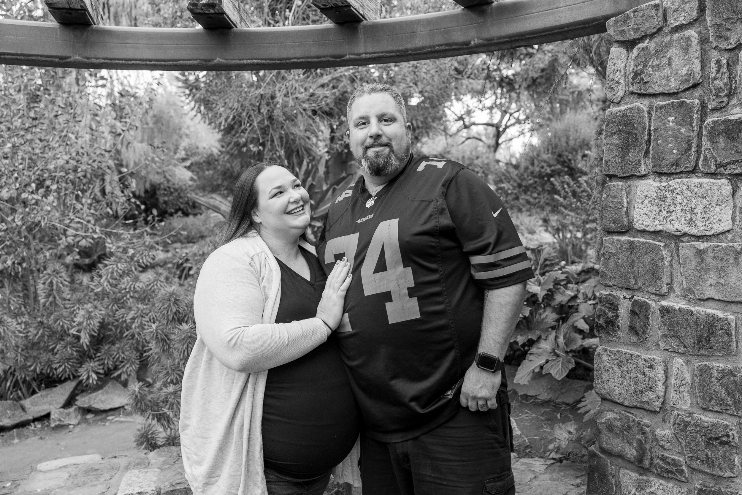 A smiling pregnant woman is touching a man in a sports jersey with the number 74, in a garden setting.