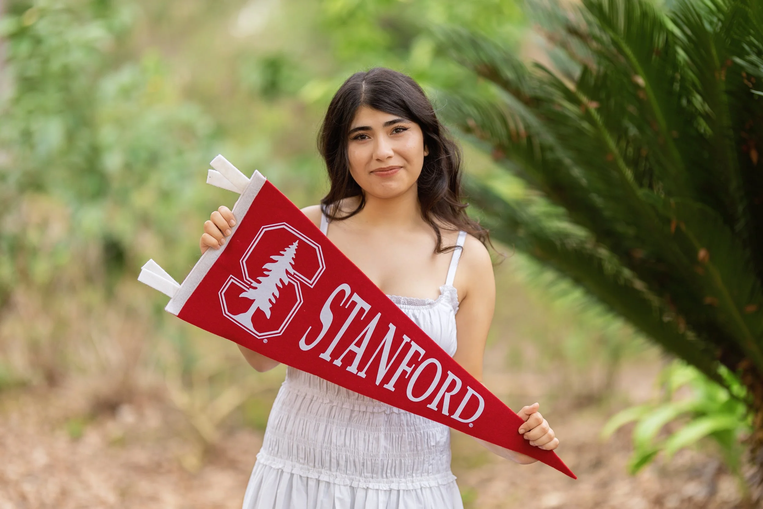 A young woman in a white dress holding a red 'Stanford' banner outdoors with green foliage in the background.