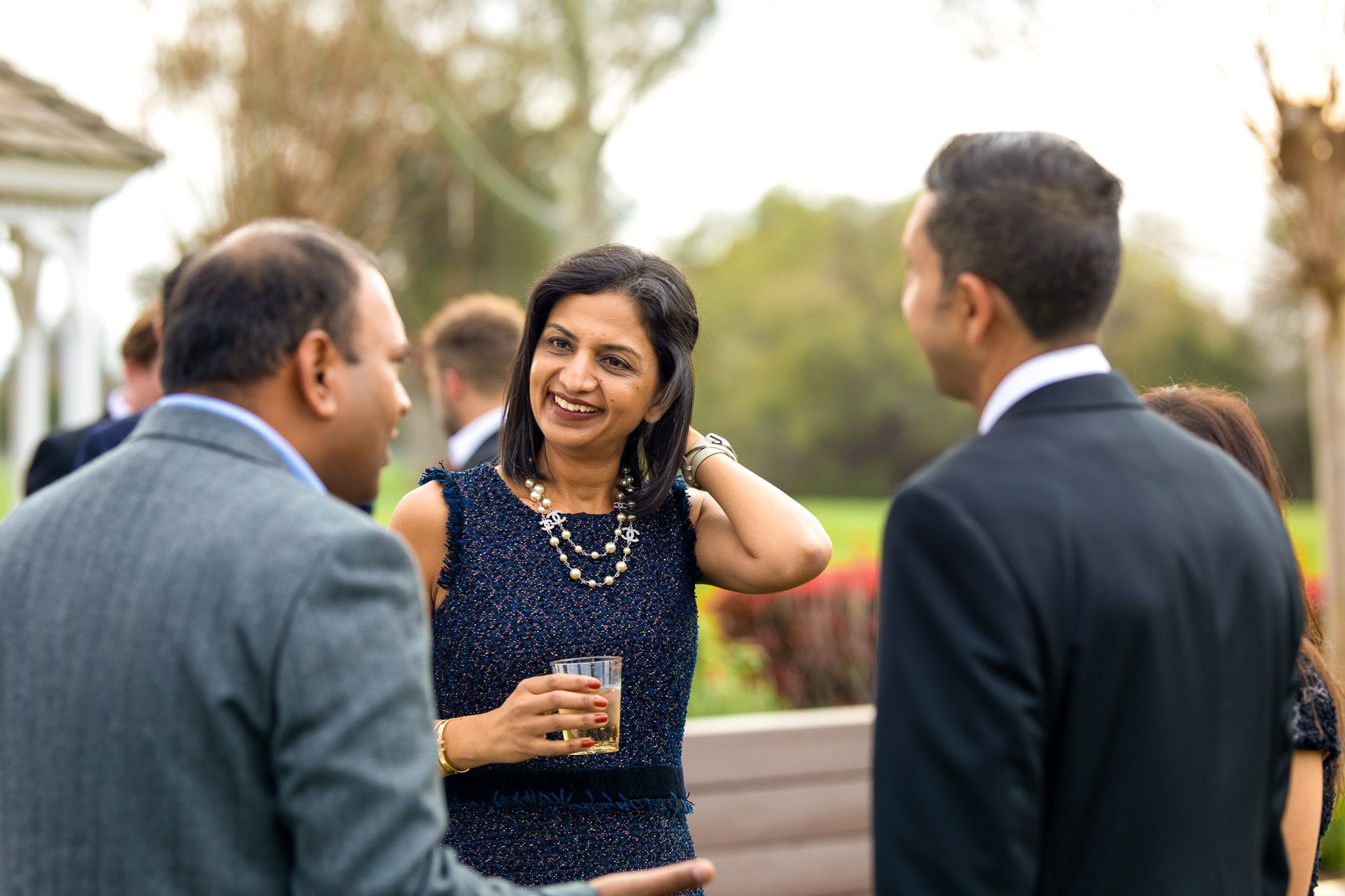 Three people engaged in conversation at an outdoor event, with a woman in a blue dress holding a drink smiling, and two men in suits talking to her, during daytime.