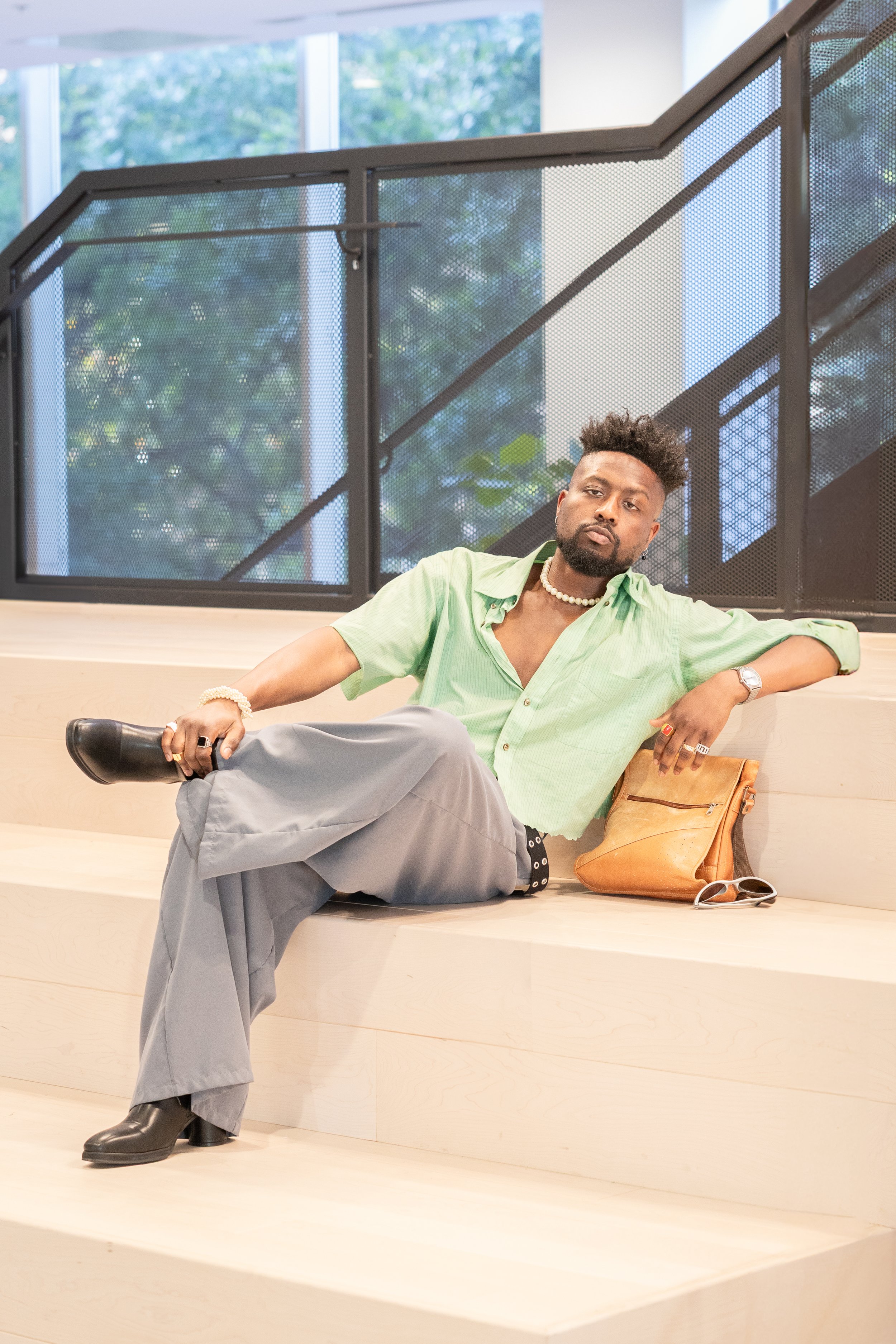 A man with dark curly hair and a beard sits on light-colored stairs, leaning back with one arm resting on his knee. He's wearing a light green short-sleeved shirt, gray wide-leg pants, and black shoes. He accessorizes with a pearl necklace, a watch,