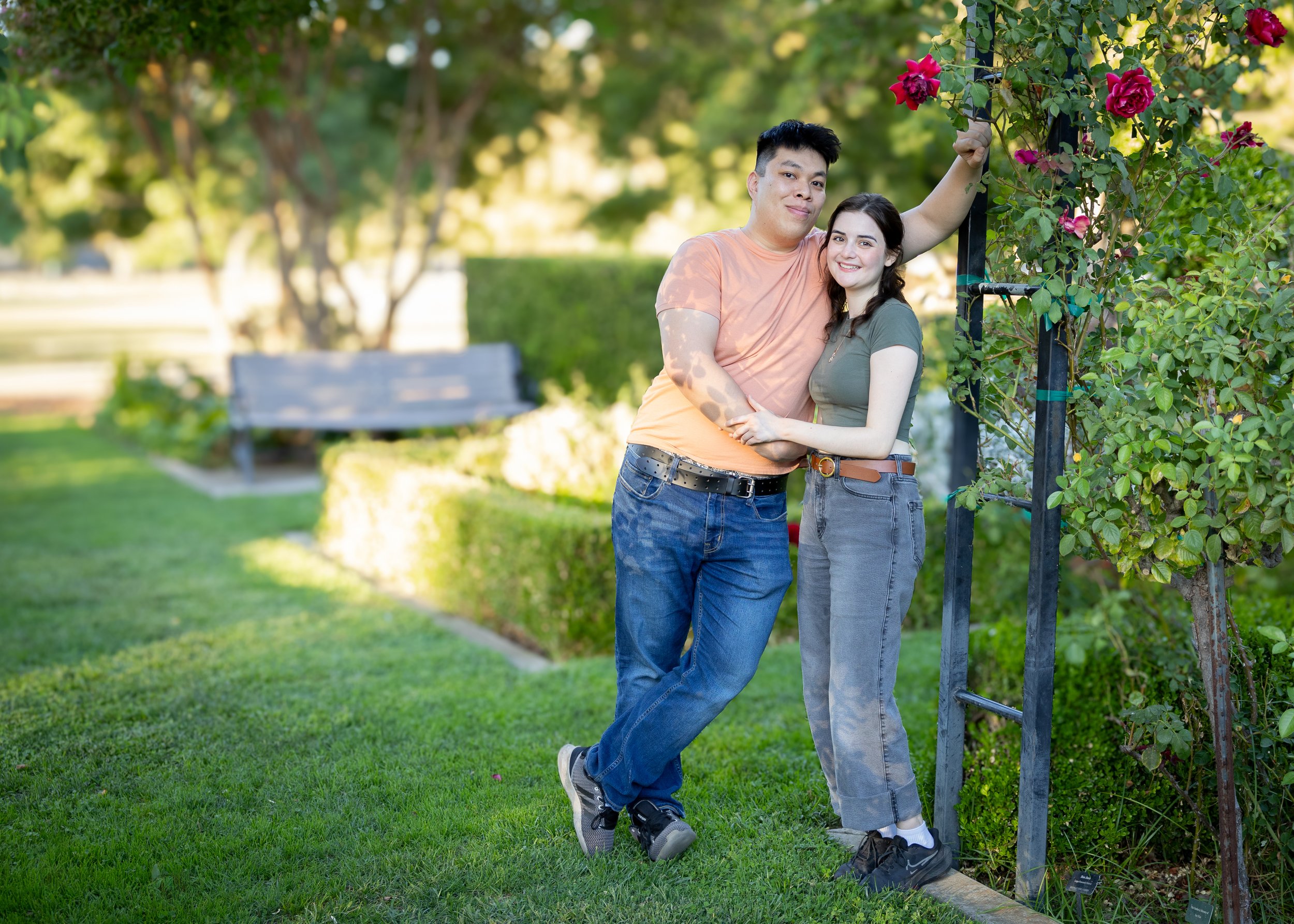 A young couple standing outdoors in a park, smiling and hugging, with greenery, a bench, and flowering plants in the background.