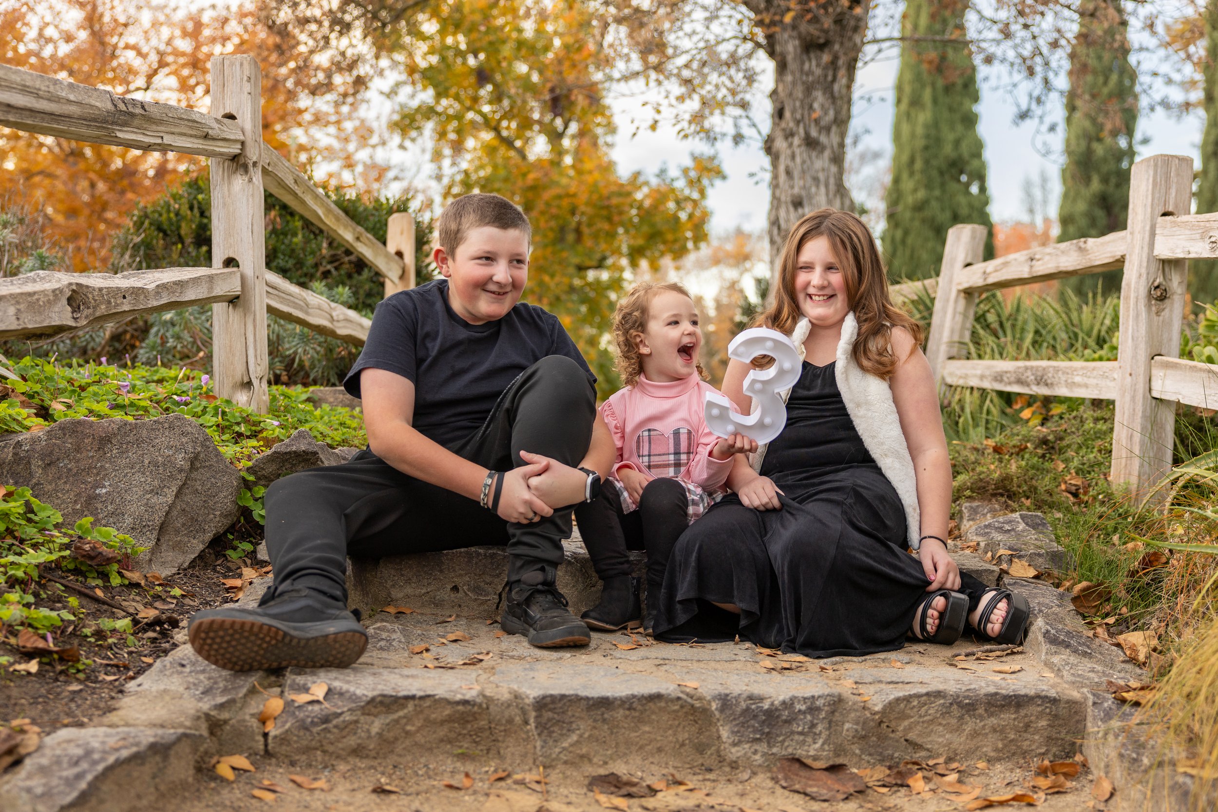 Three children sitting on stone steps outdoors during autumn, smiling and laughing, with one holding a large decorative number three.