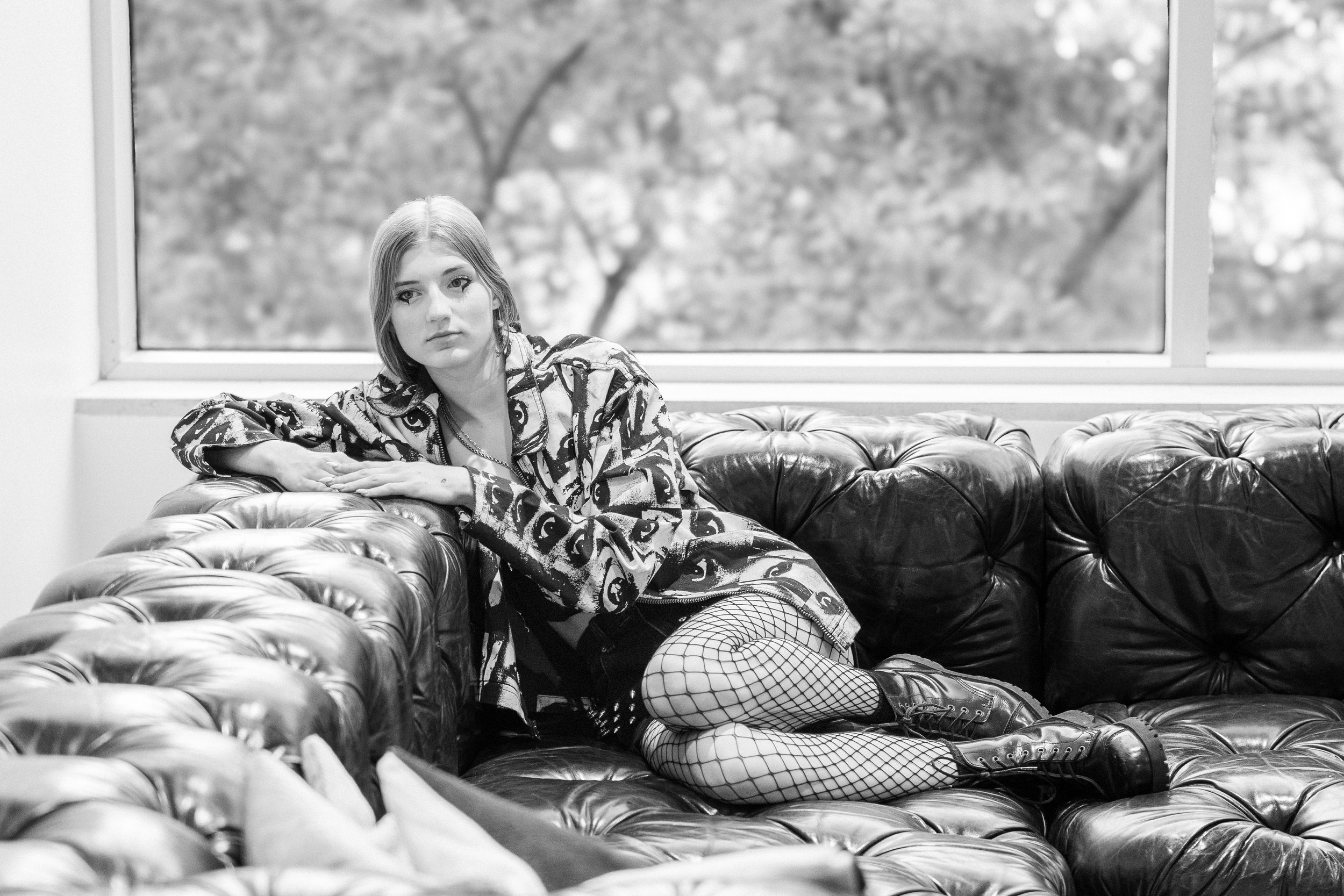 A young woman with long hair lies on a leather tufted couch, resting her head on her arm, wearing a patterned jacket, fishnet stockings, and combat boots, in a room with large window and outdoor trees in the background.