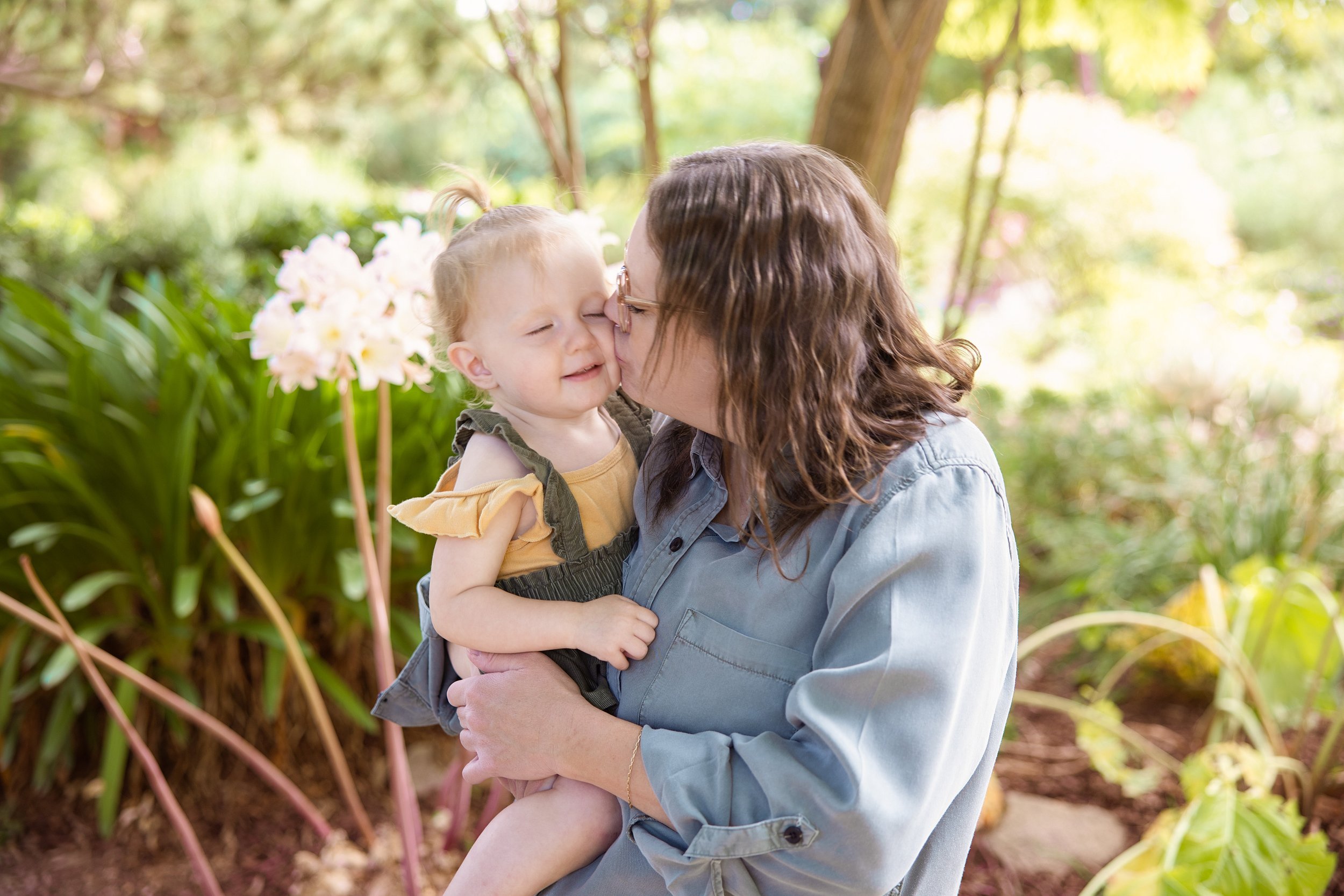 A woman with brown hair and glasses is holding a young girl with blonde hair in a ponytail. They are outdoors in a garden with green plants and trees, and the woman is kissing the girl's cheek while the girl smiles with eyes closed.