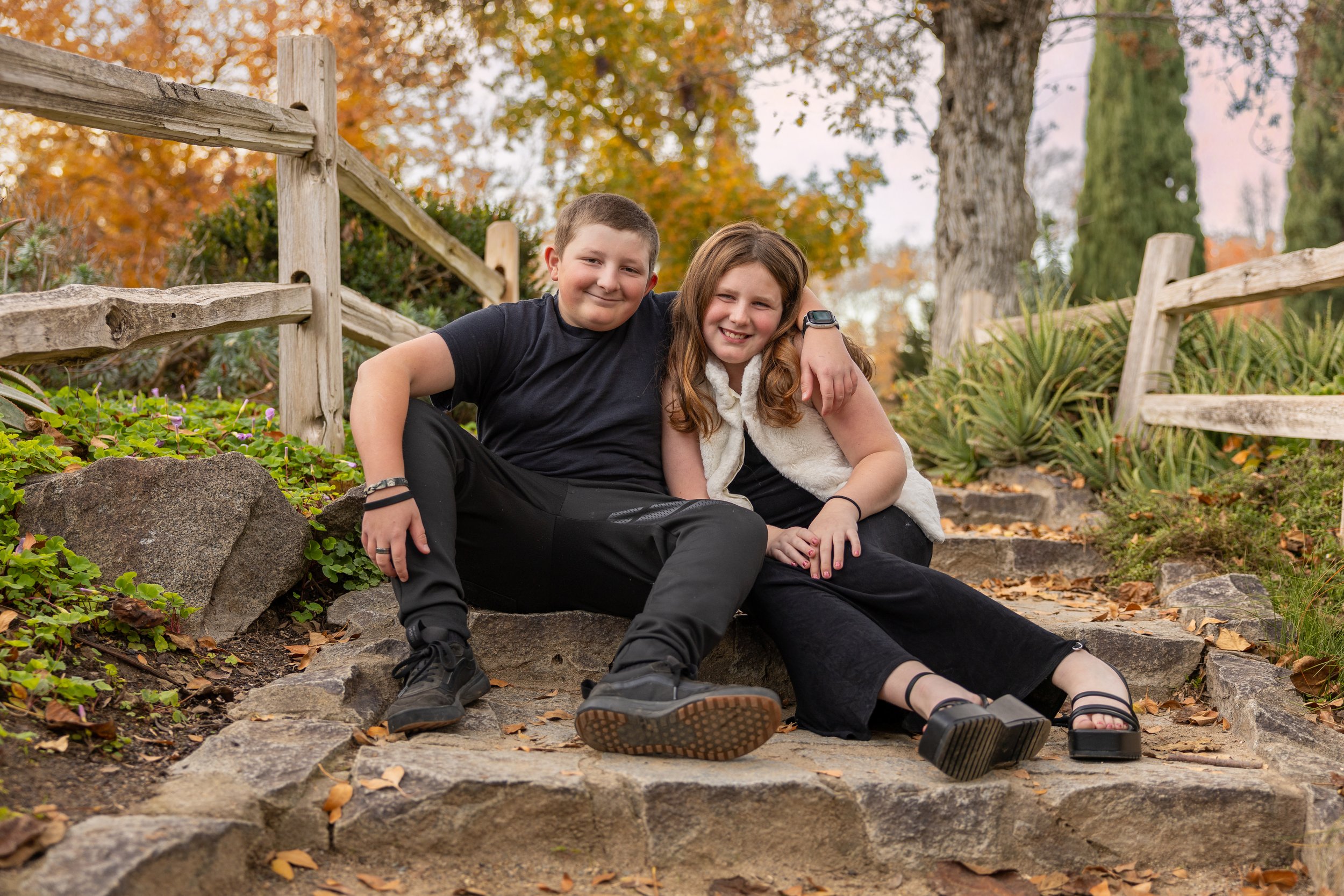 Two children sitting on stone steps outdoors during autumn, smiling with their arms around each other, surrounded by fall foliage and a wooden fence.