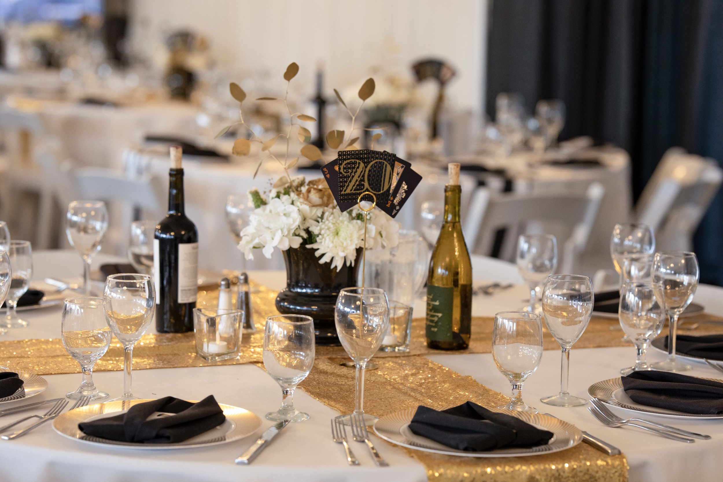 Elegant banquet table decorated with black napkins, gold table runner, floral centerpiece, wine bottles, and glasses, set for a celebration with a table card displaying the number '20'.