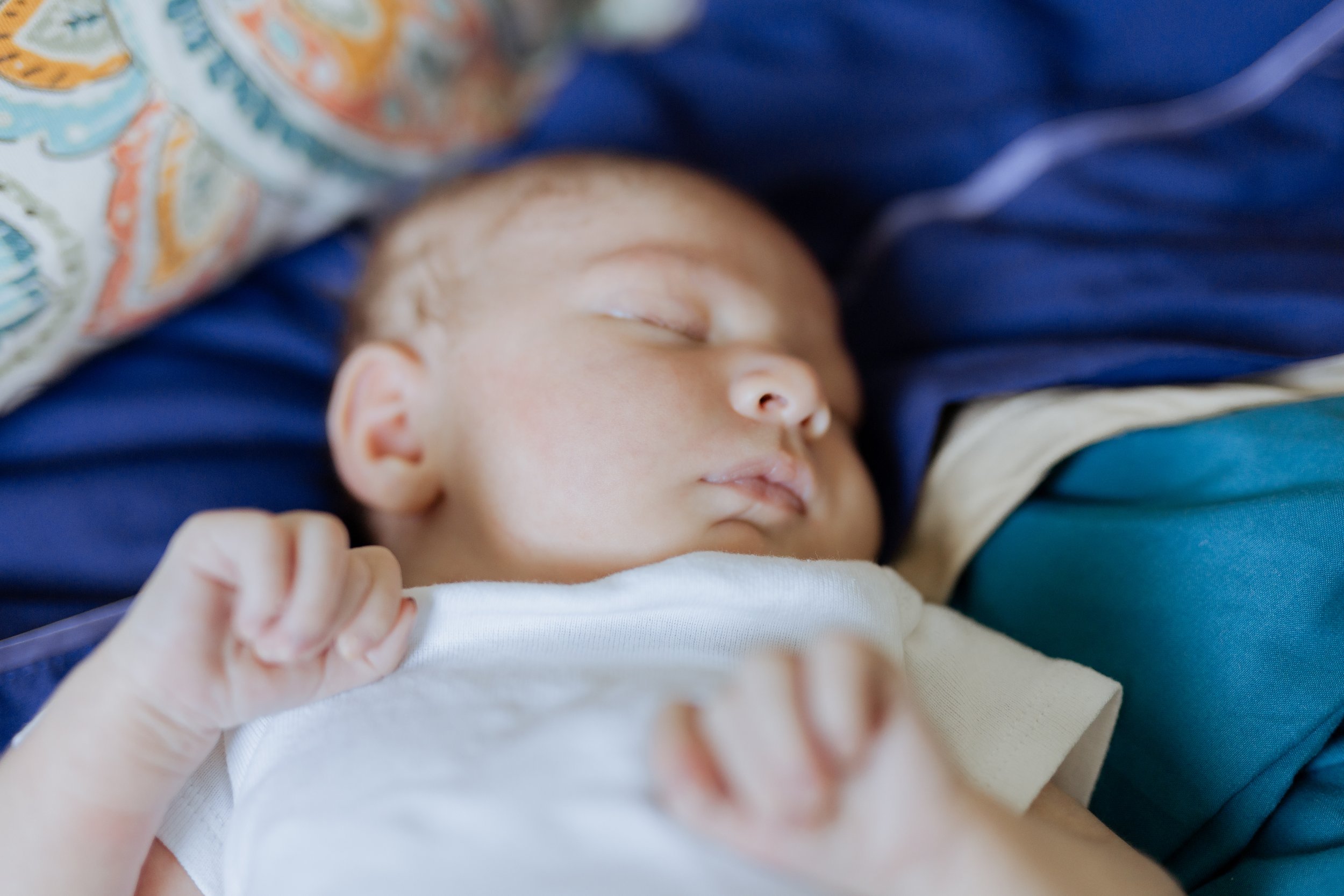 A young child with light skin sleeping peacefully with a white shirt, lying on a blue pillow and surrounded by colorful patterned fabric.