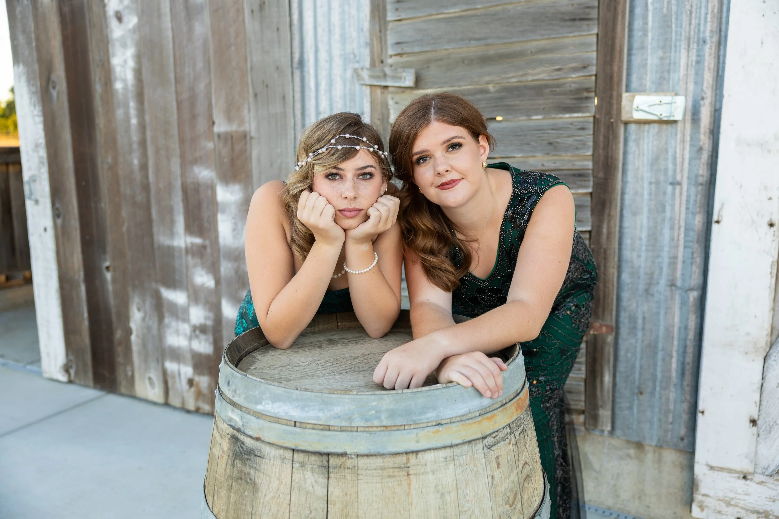 Two young women leaning on an old wooden barrel, posing in front of a rustic wooden wall outdoors.