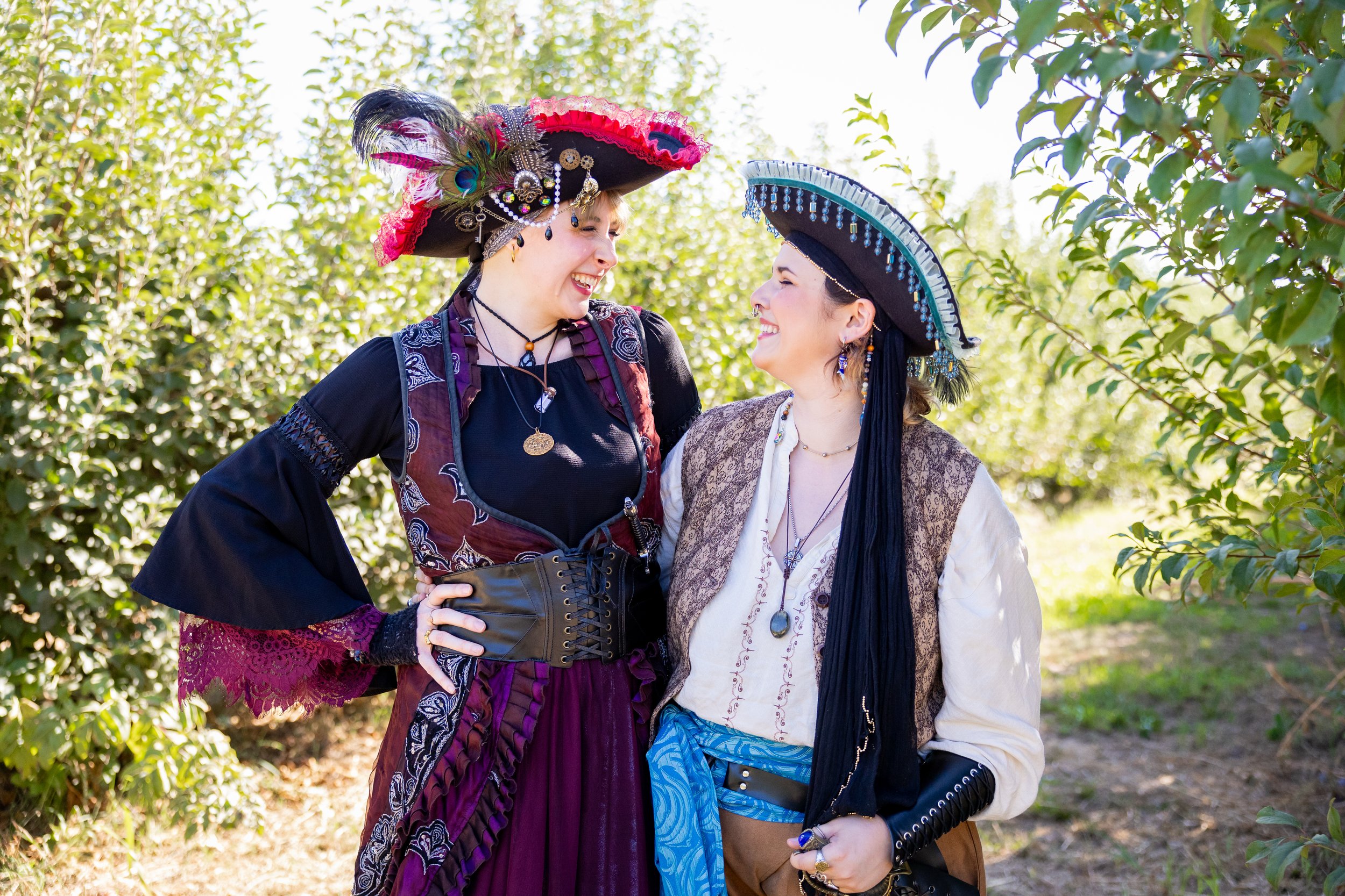 Two women dressed as pirates standing outdoors, smiling and looking at each other, with trees in the background.