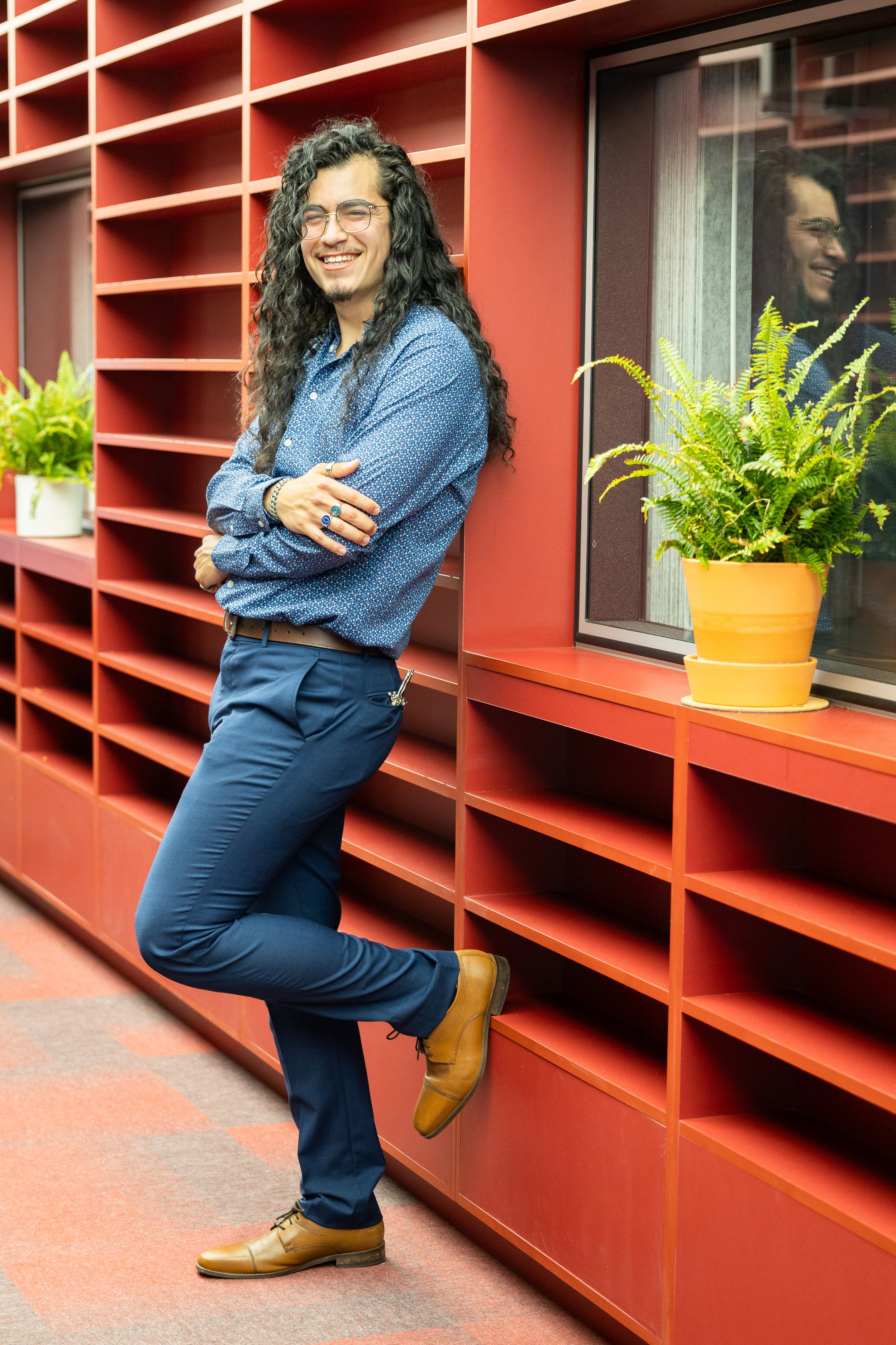 A man with long curly dark hair, glasses, and a beard, standing and leaning against a red bookshelf in an office setting. He is smiling, wearing a blue patterned shirt, dark blue pants, and tan shoes. There are potted plants on the shelf beside him a