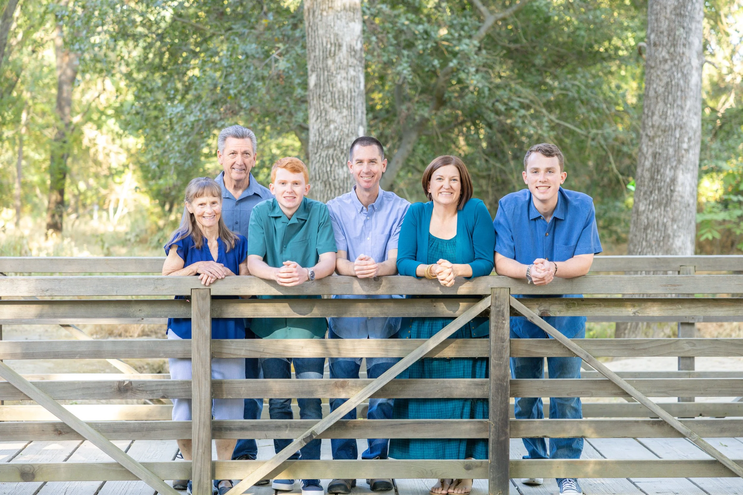 A multi-generational family of seven posing outdoors behind a wooden fence, surrounded by trees and greenery.