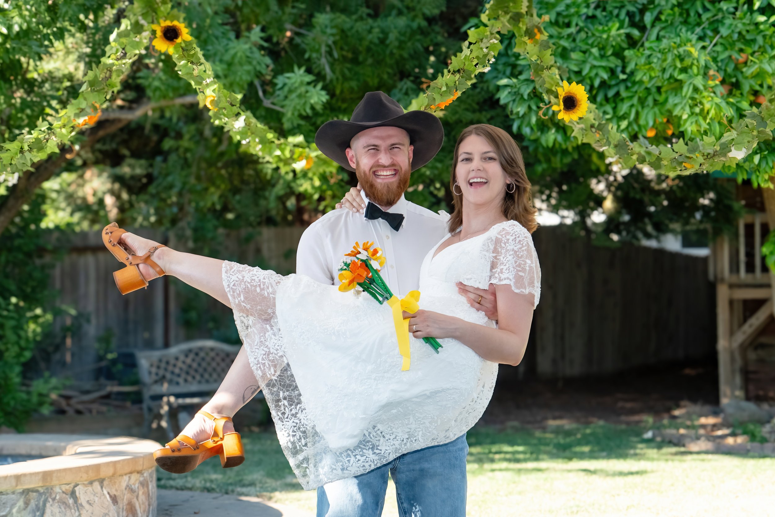 Couple in wedding attire with the man wearing a cowboy hat, holding the woman in a white lace dress, outside under a tree with yellow sunflowers, both smiling and happy.